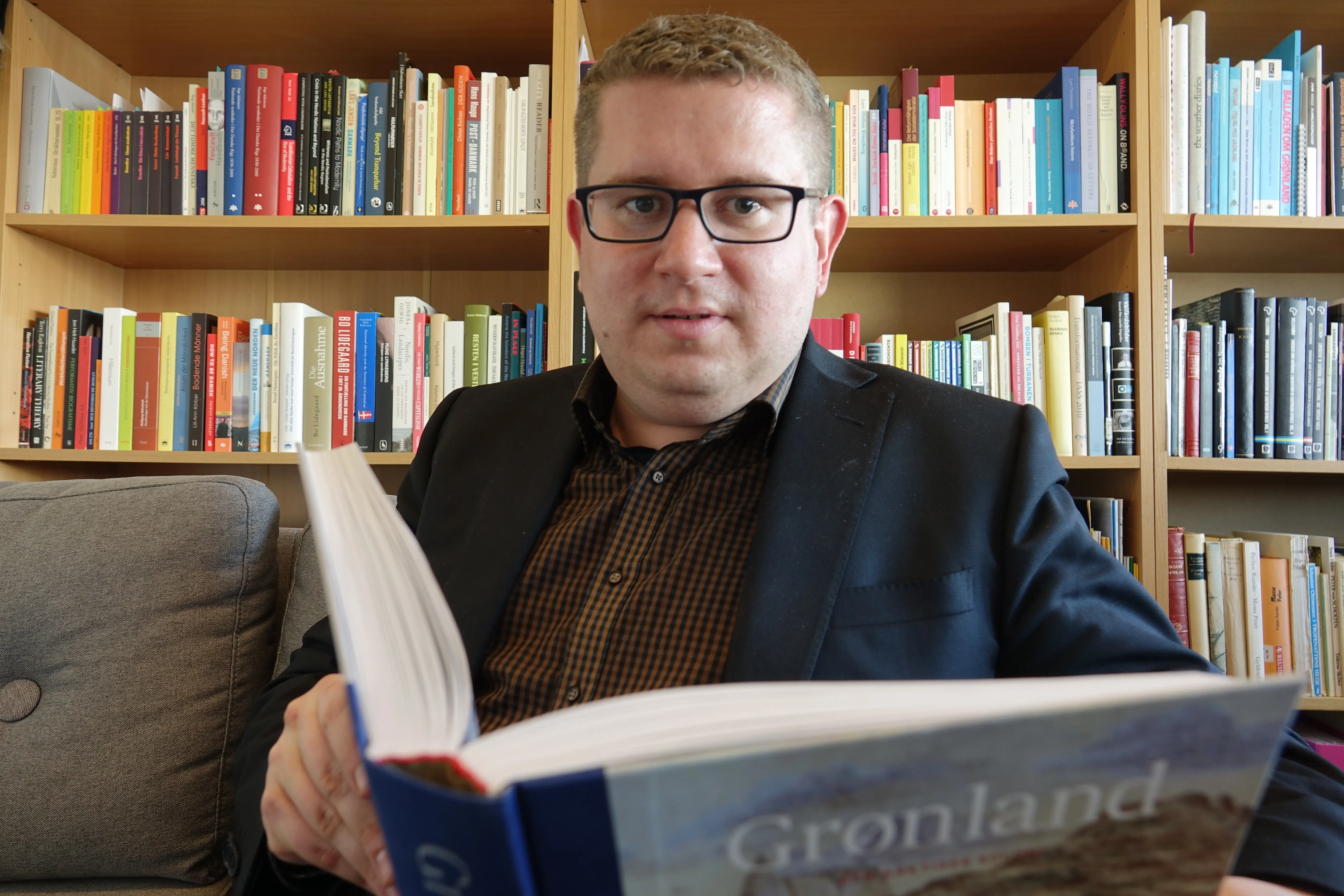 A man holds a book reading Gronland in a library sitting on a couch
