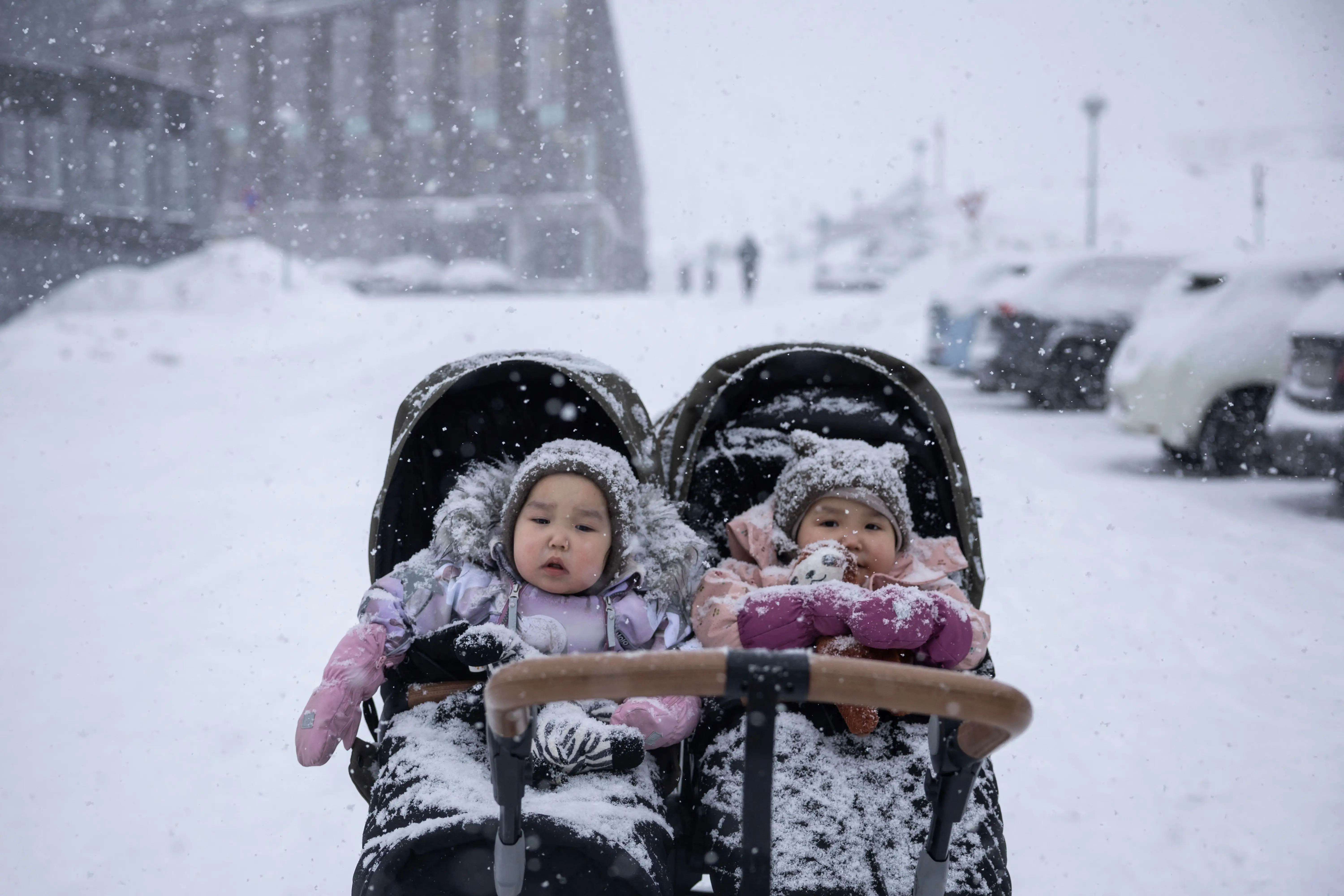 Two babies in a double stroller in the snow with cars in the background