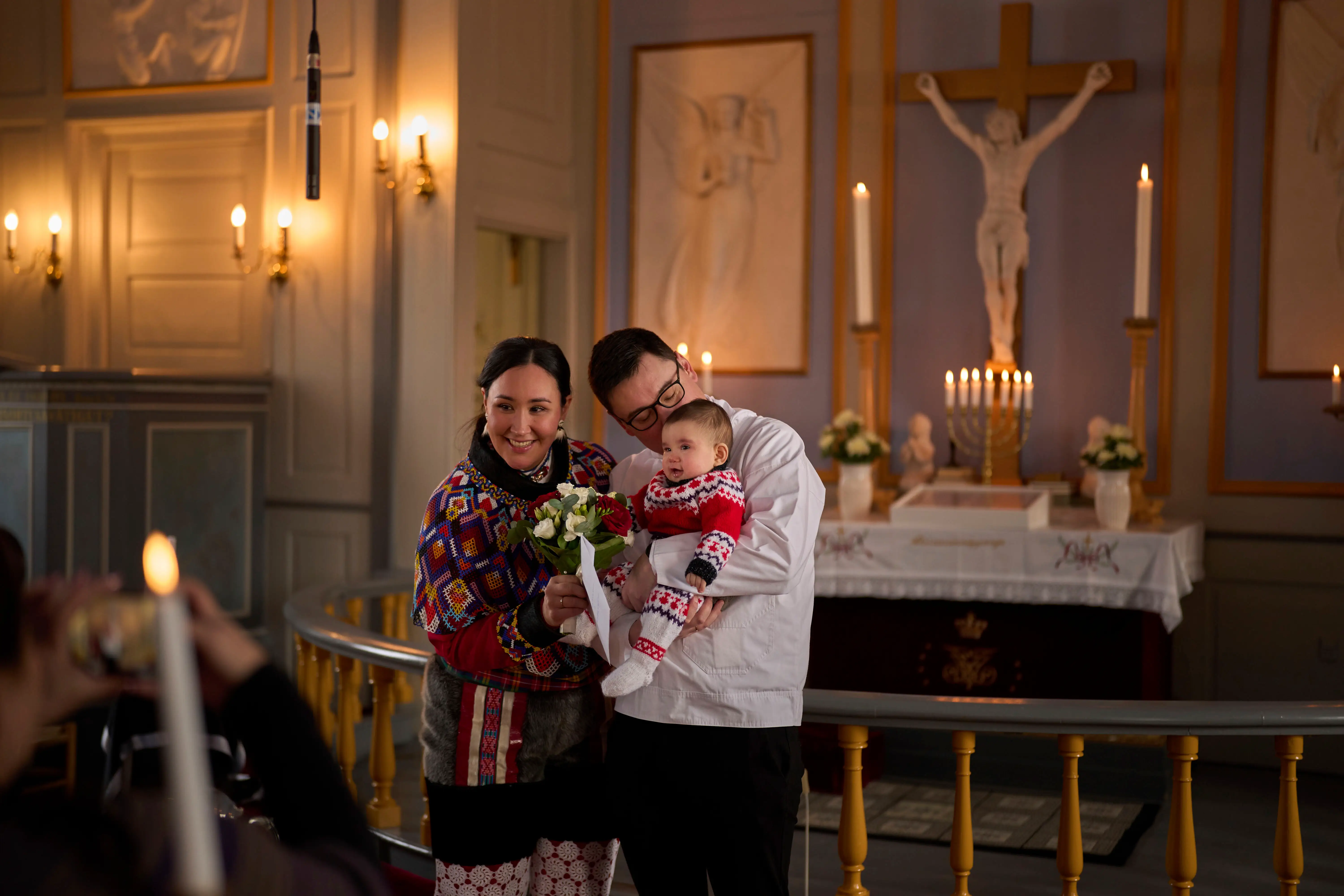 A couple holds their baby in a church in front of a crucifix and candles on an alter