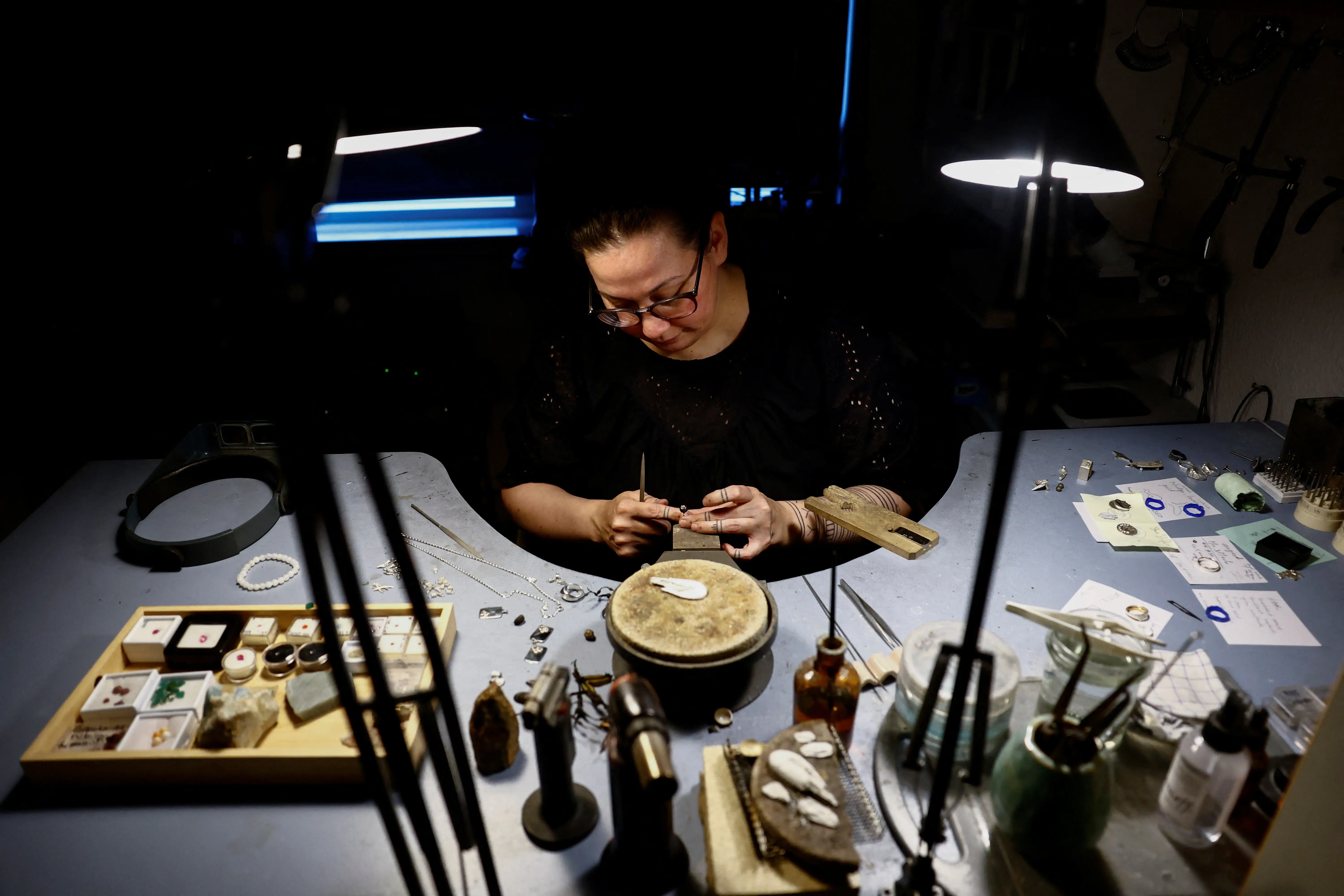 A woman works on a piece of jewelry with tools and lamps covering her desk