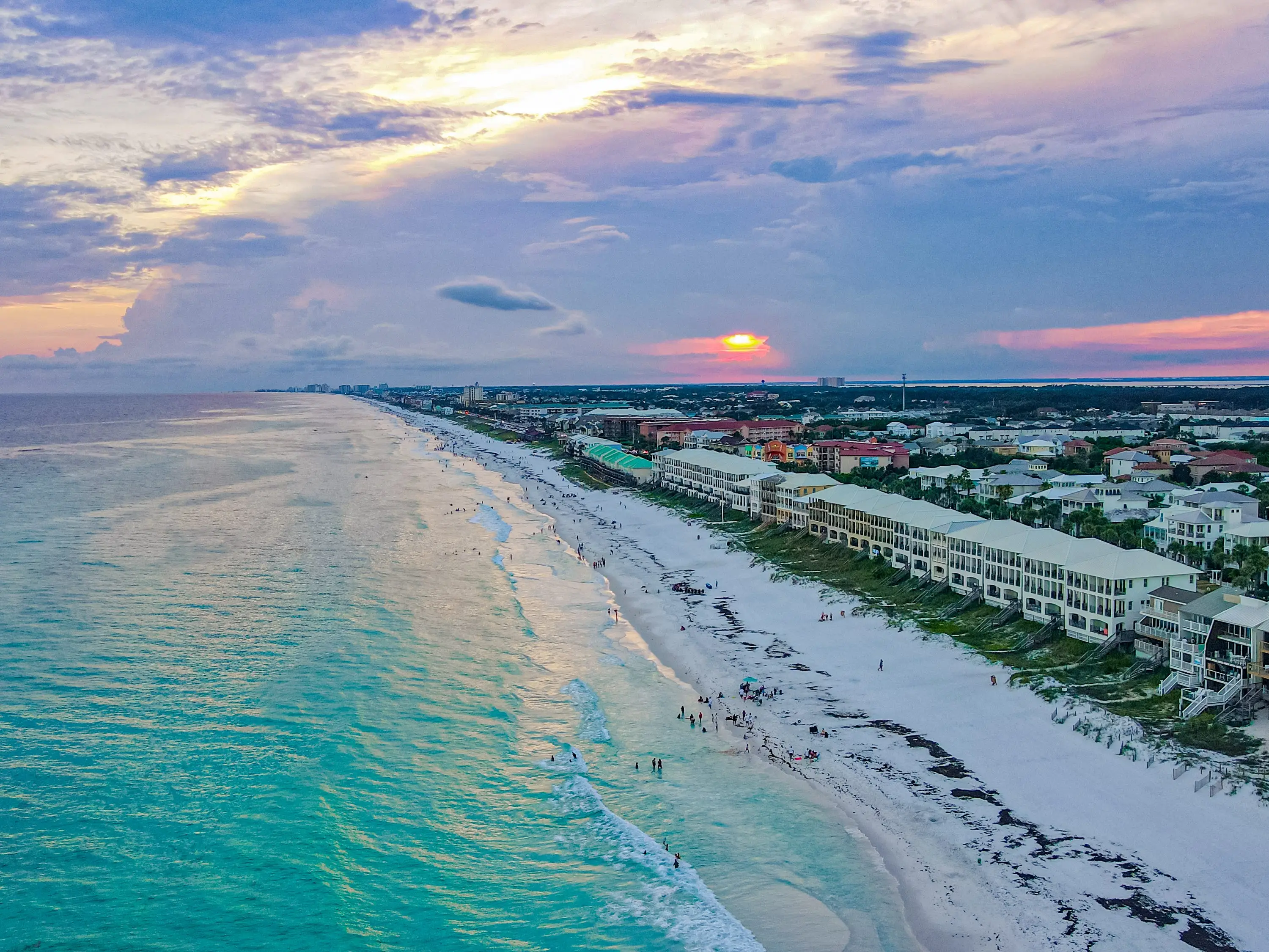 An aerial view of a beach at sunset, with houses along the dunes.