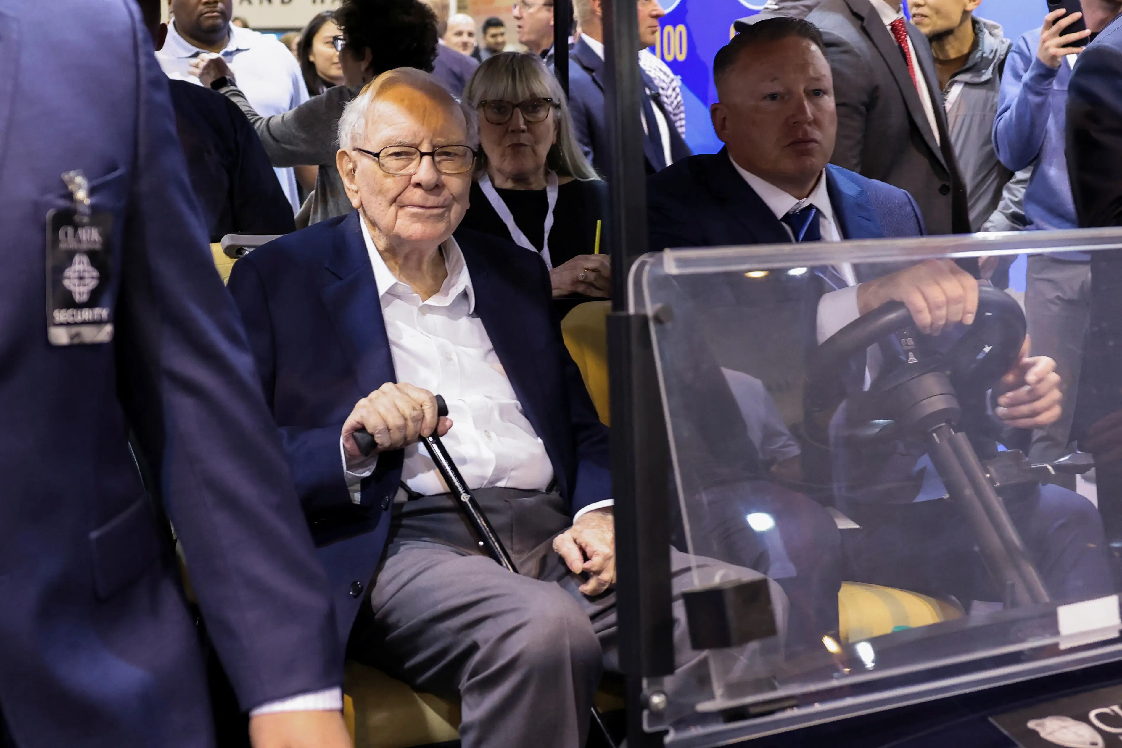 Warren Buffett in a suit in a golf car surrounded by people.
