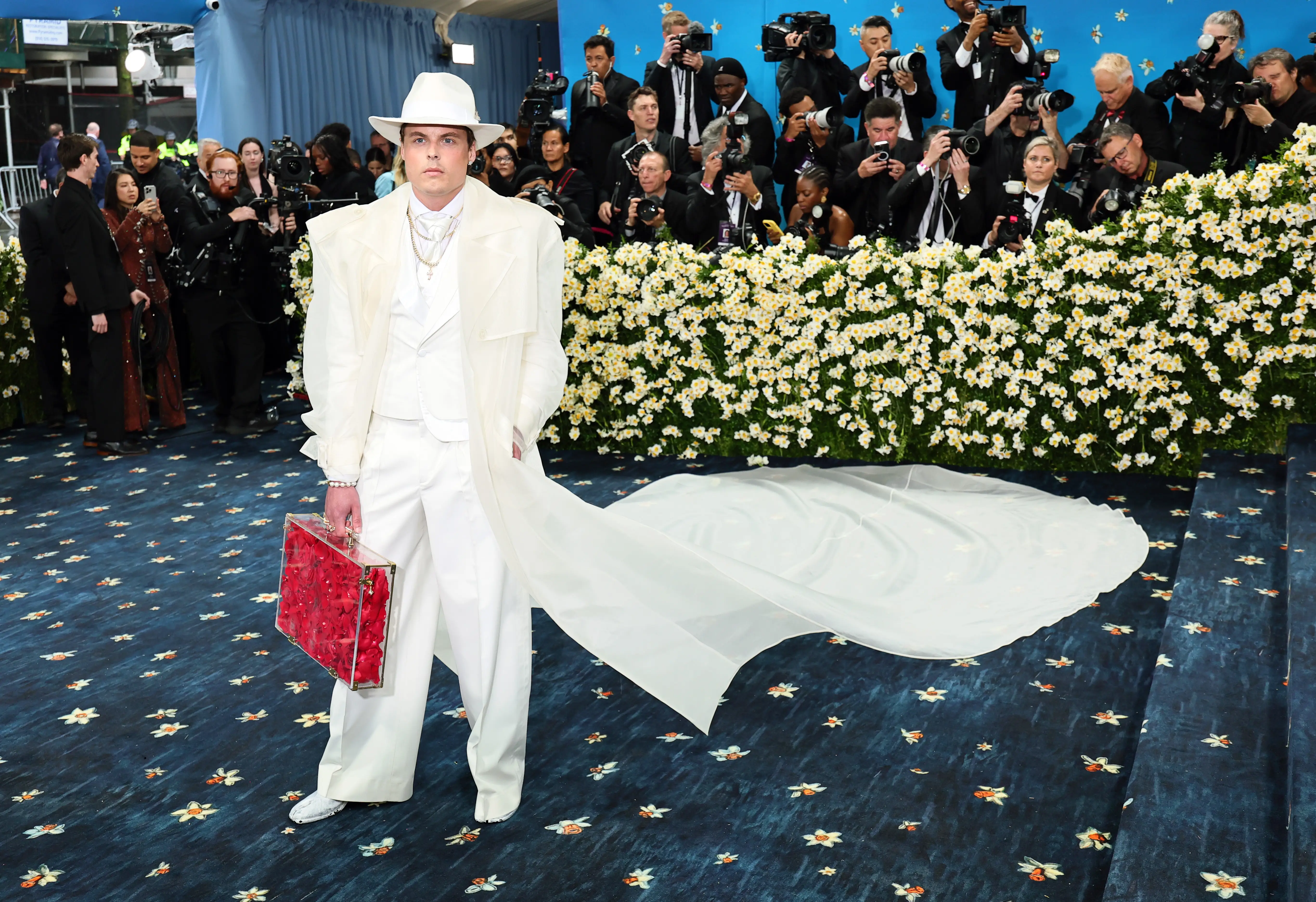 Gustav Magnar Witzøe on the navy carpet at the Met Gala. He's wearing a white suit with a big flowing white cap and a white cowboy hat. He's also holding a clear briefcase full of red flowers.