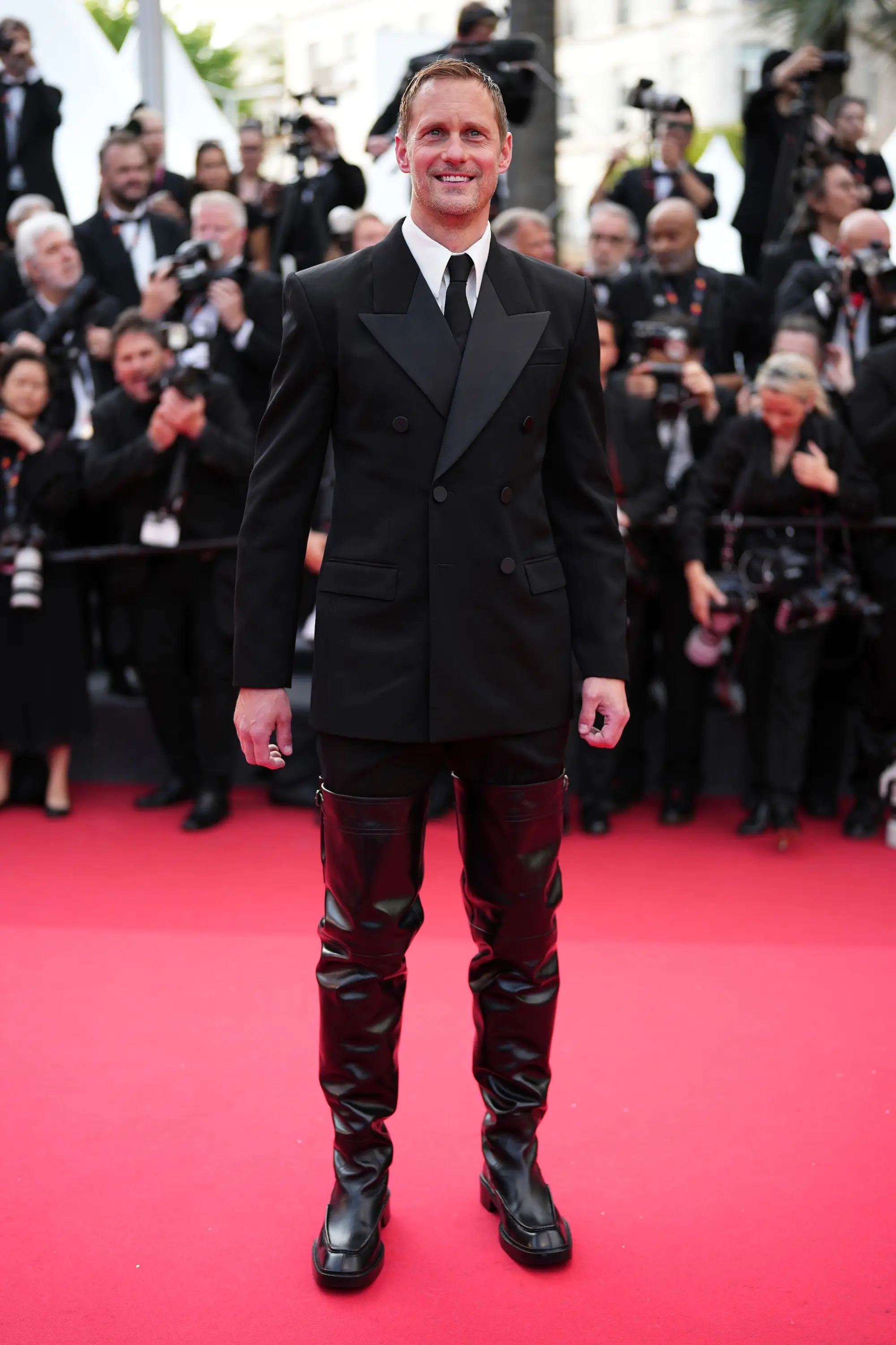 Alexander Skarsgard wears a suit and thigh-high leather boots on the Cannes Film Festival red carpets, with photographers behind him.