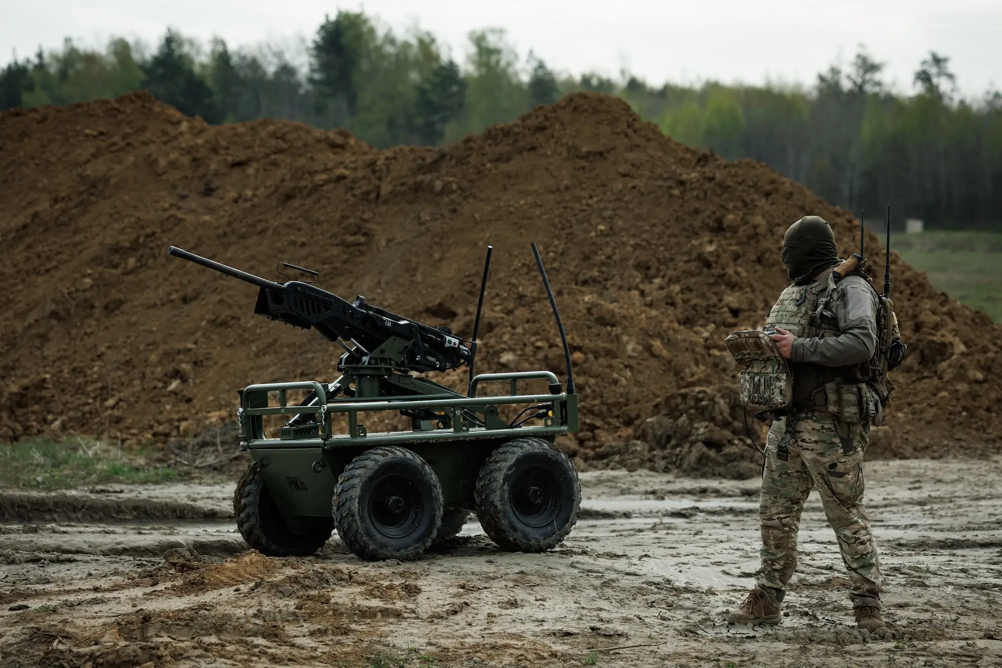 A man in camouflage gear stands looking at a large wheeled robot with a machine gun on top in front of a large mound of brown earth