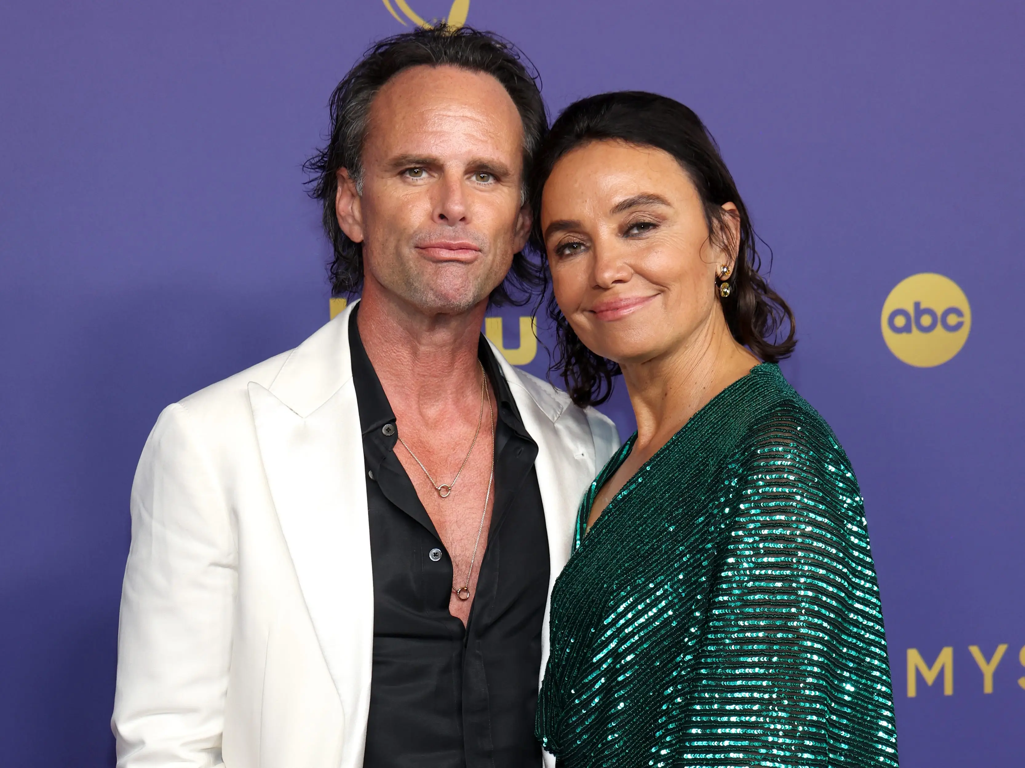 A man and a woman at an event. On the left, the man has long swept-back black hair. He's wearing a white blazer over an open-collared black shirt and black trousers. On the right, the woman also has her black hair swept back, and is wearing a glittery green dress. They're standing against a purple backdrop with gold logos for Hulu, ABC, and the Emmys on it.