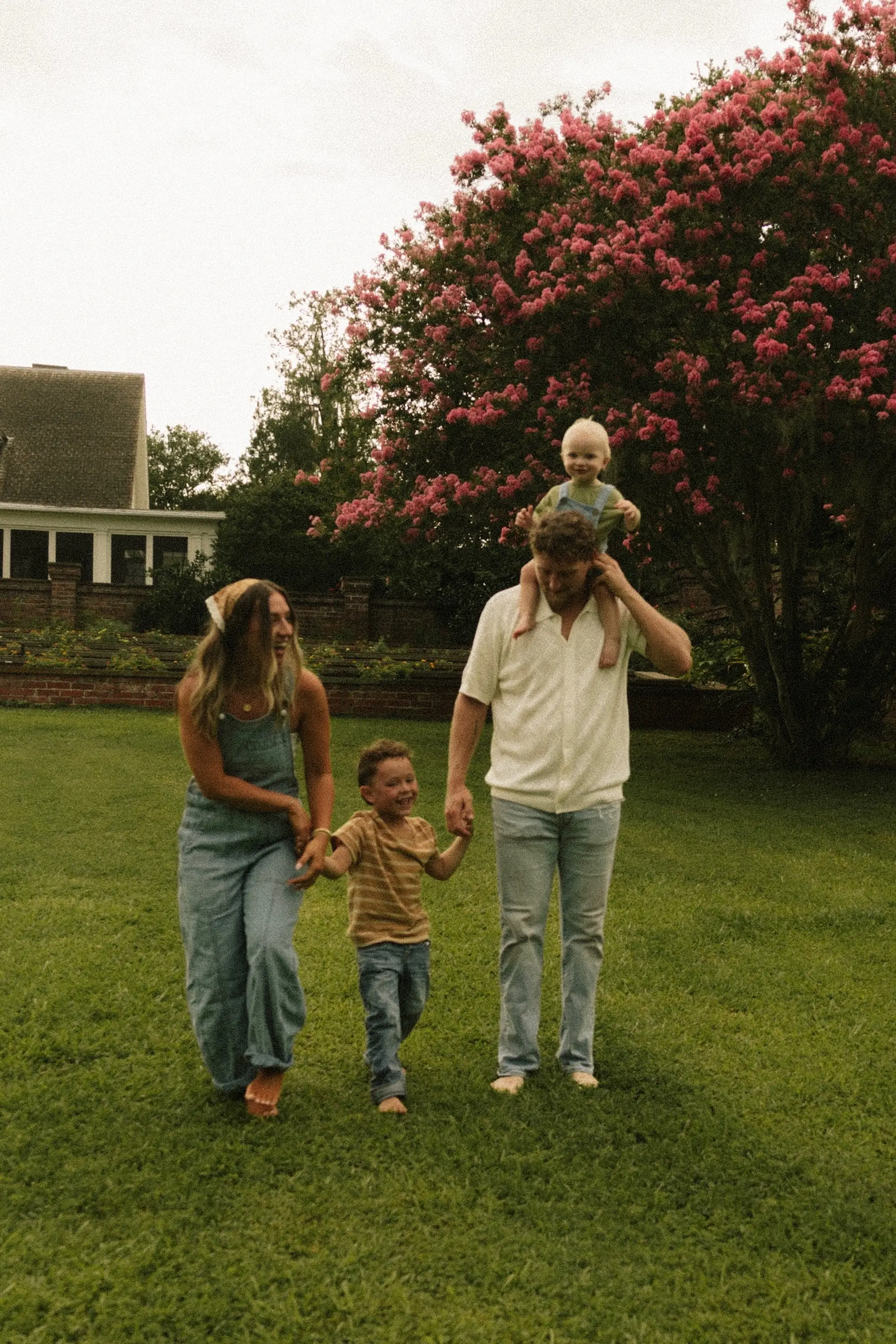 A woman and man hold hand with a little boy in a field. The man has another little boy on his shoulders.