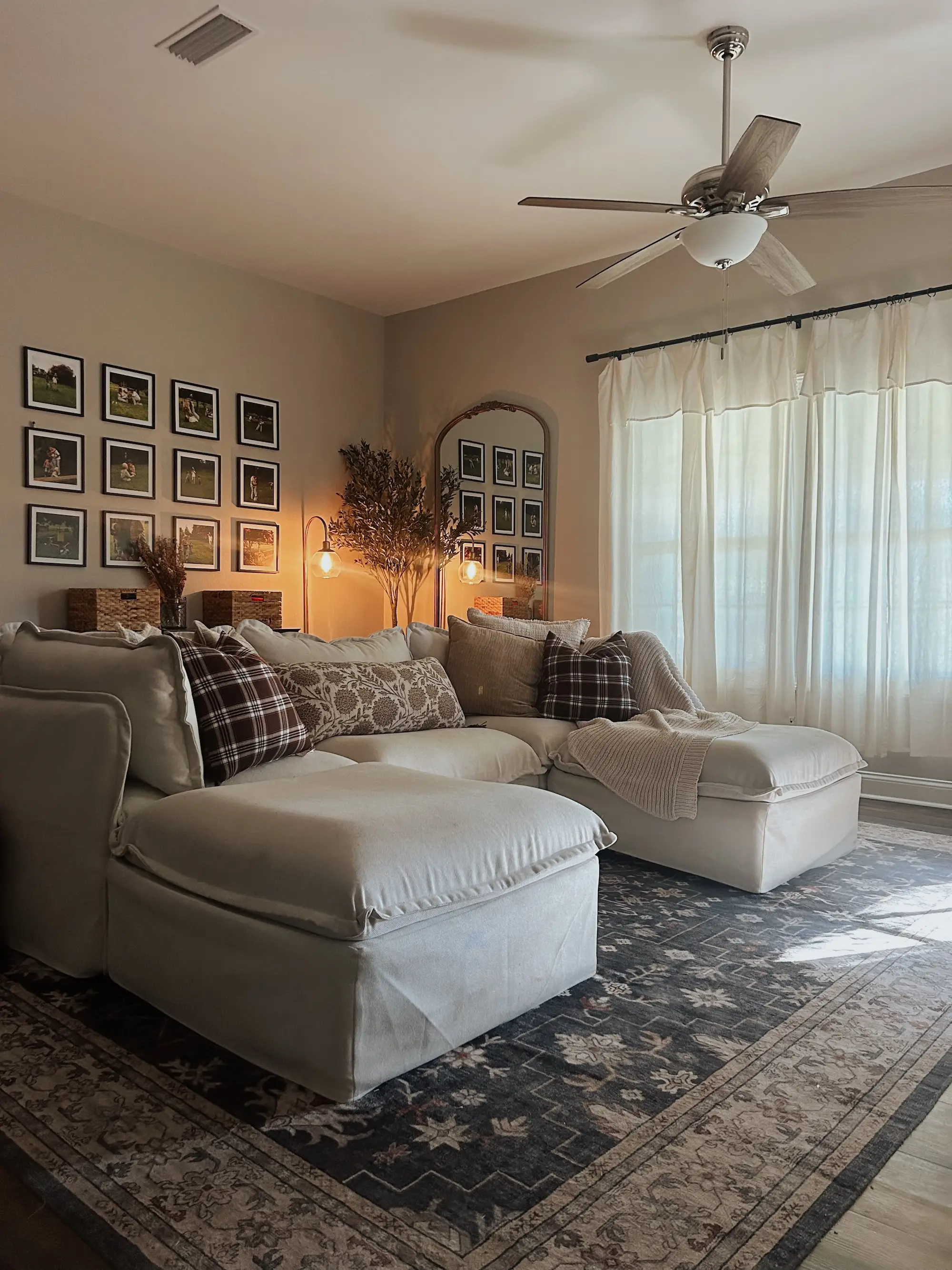 A living room with a large cream couch and a floral rug. A gallery wall of photos sits on the back wall.