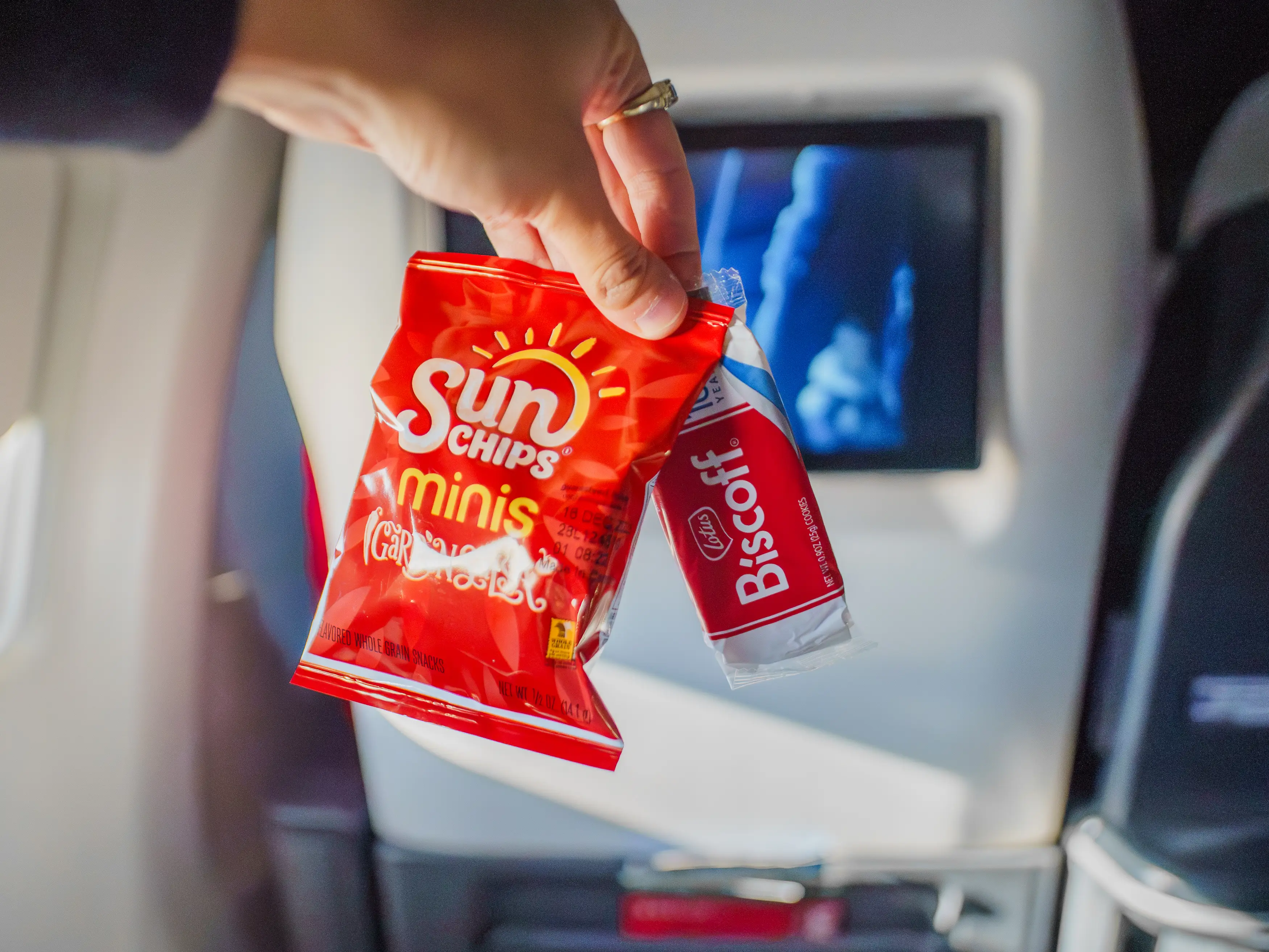 A hand holds a Biscoff cookie package and a small bag of Sun Chips in front of a first-class seat-back entertainment screen