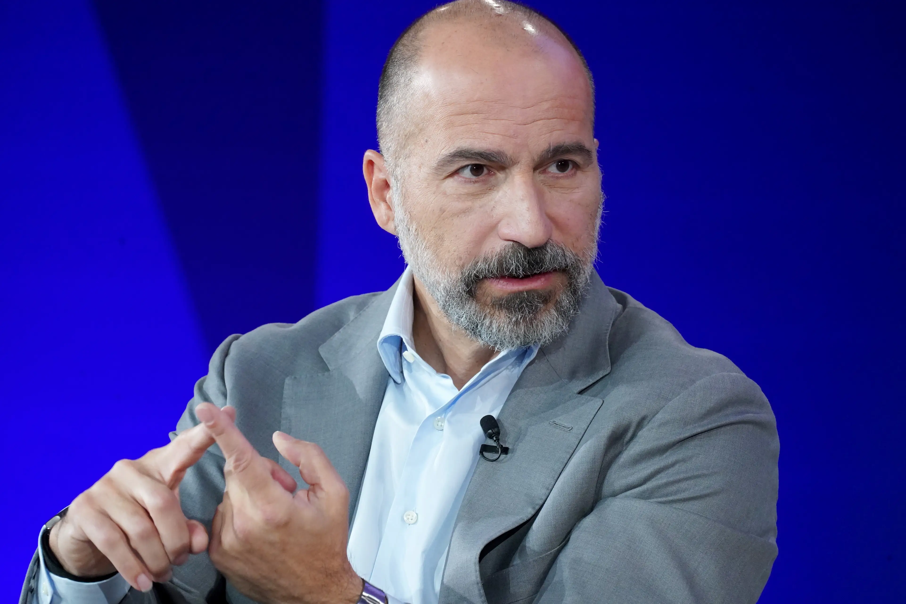 Uber CEO Dara Khosrowshahi speaks at a conference while wearing a light grey suit, light blue dress shirt, and crossing his two pointer fingers.