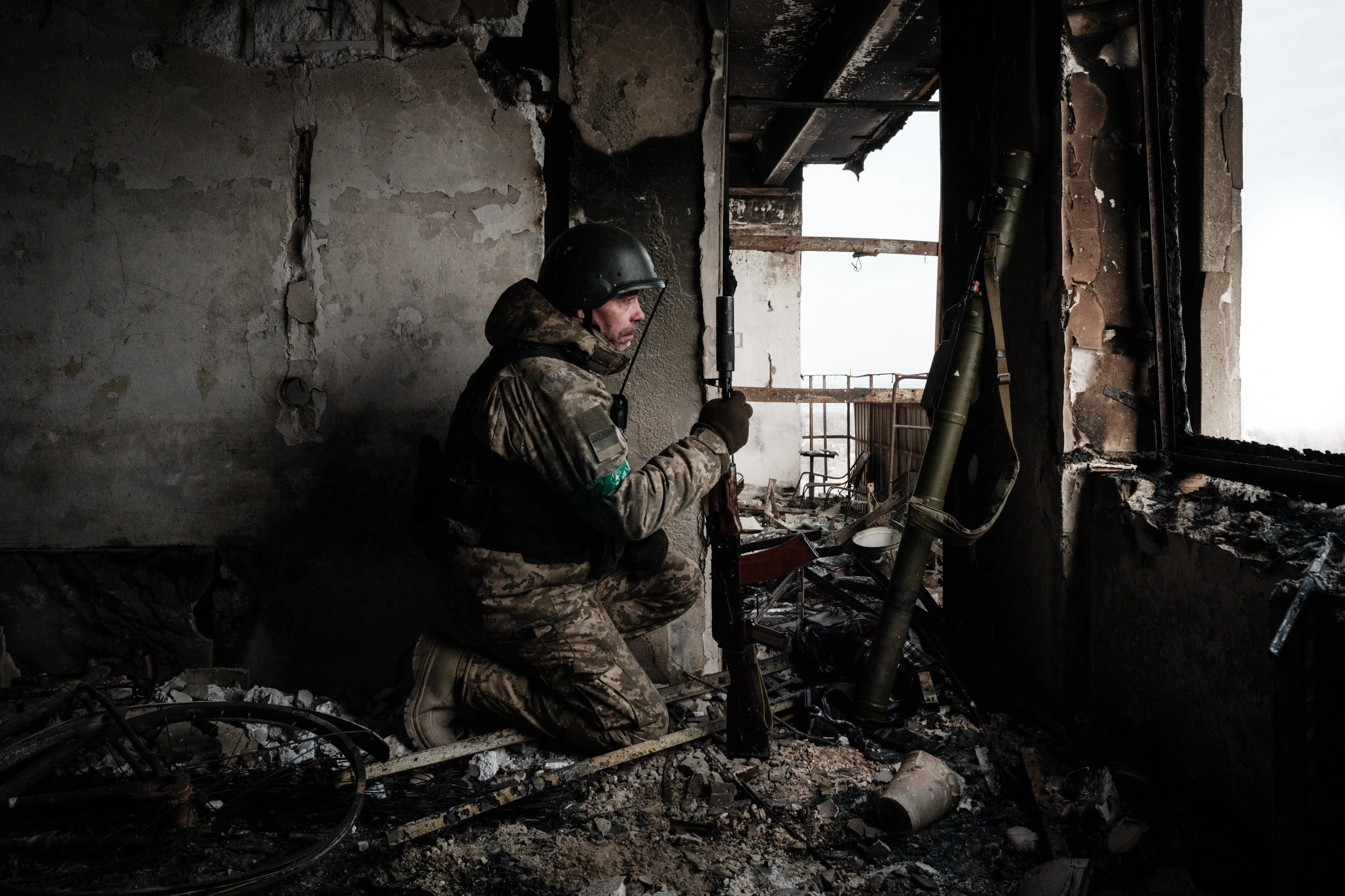 A man in camouflage gear and a helmet kneels in a rubble-filled room with destroyed walls