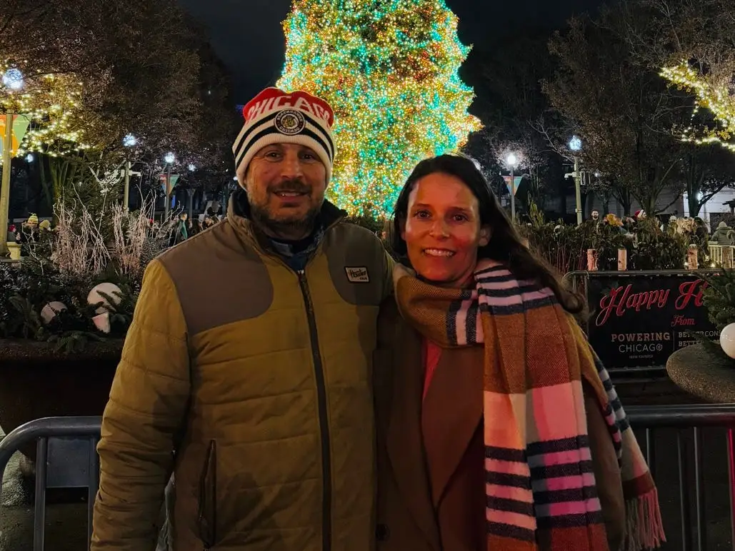 Author Ash Jurberg  and wife smiling in front of lit-up tree at night