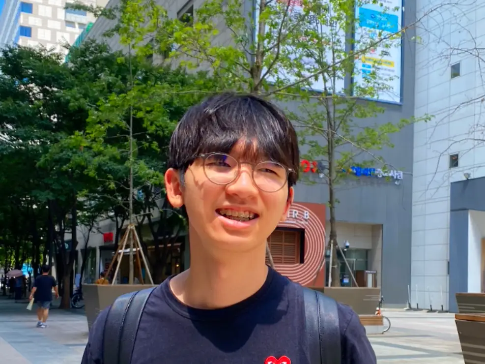 Smiling young man with glasses and a backpack standing outdoors with trees and buildings behind him.