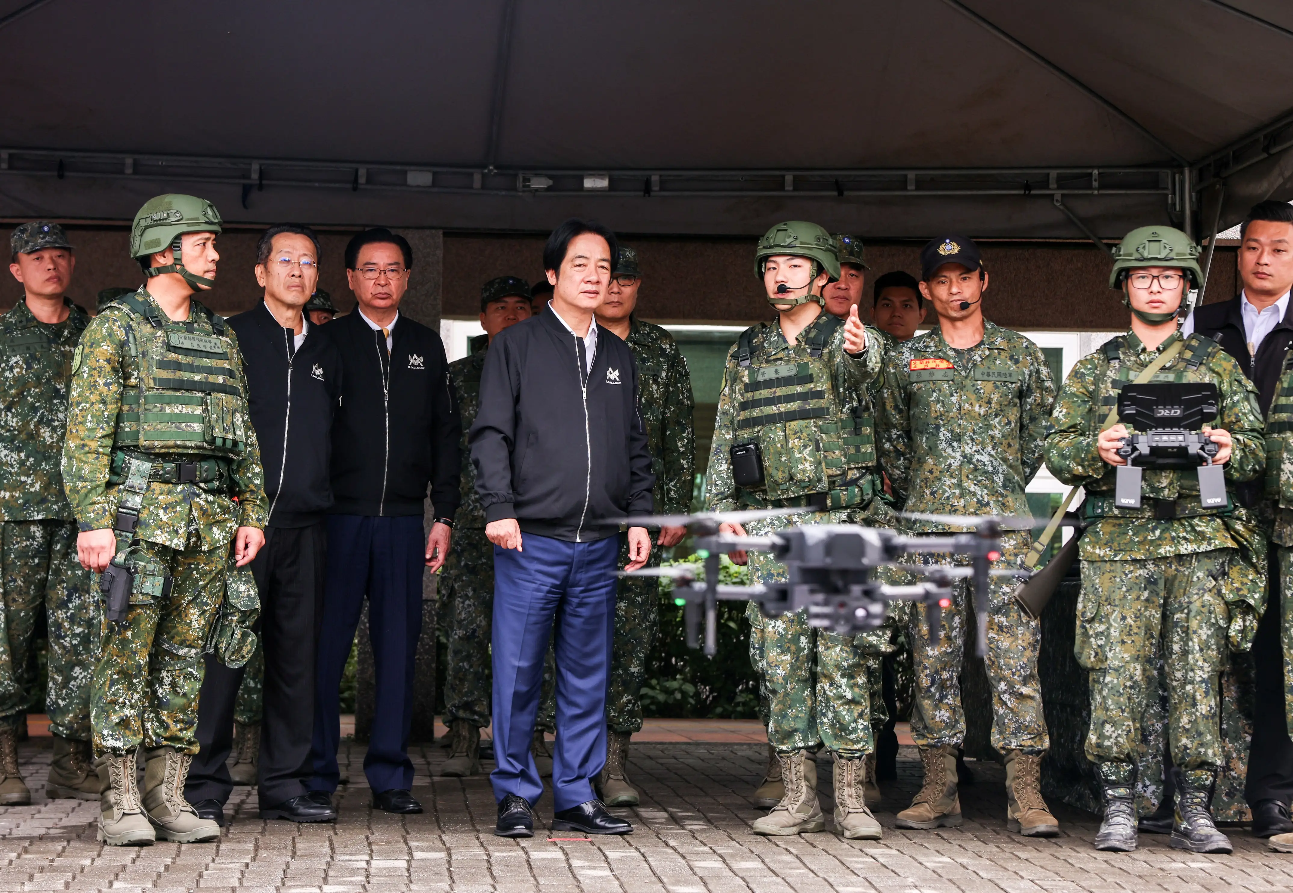 A group of Taiwanese men stand in a line under a tent. One man wearing camouflage is presenting a small grey drone, which is flying in the foreground.