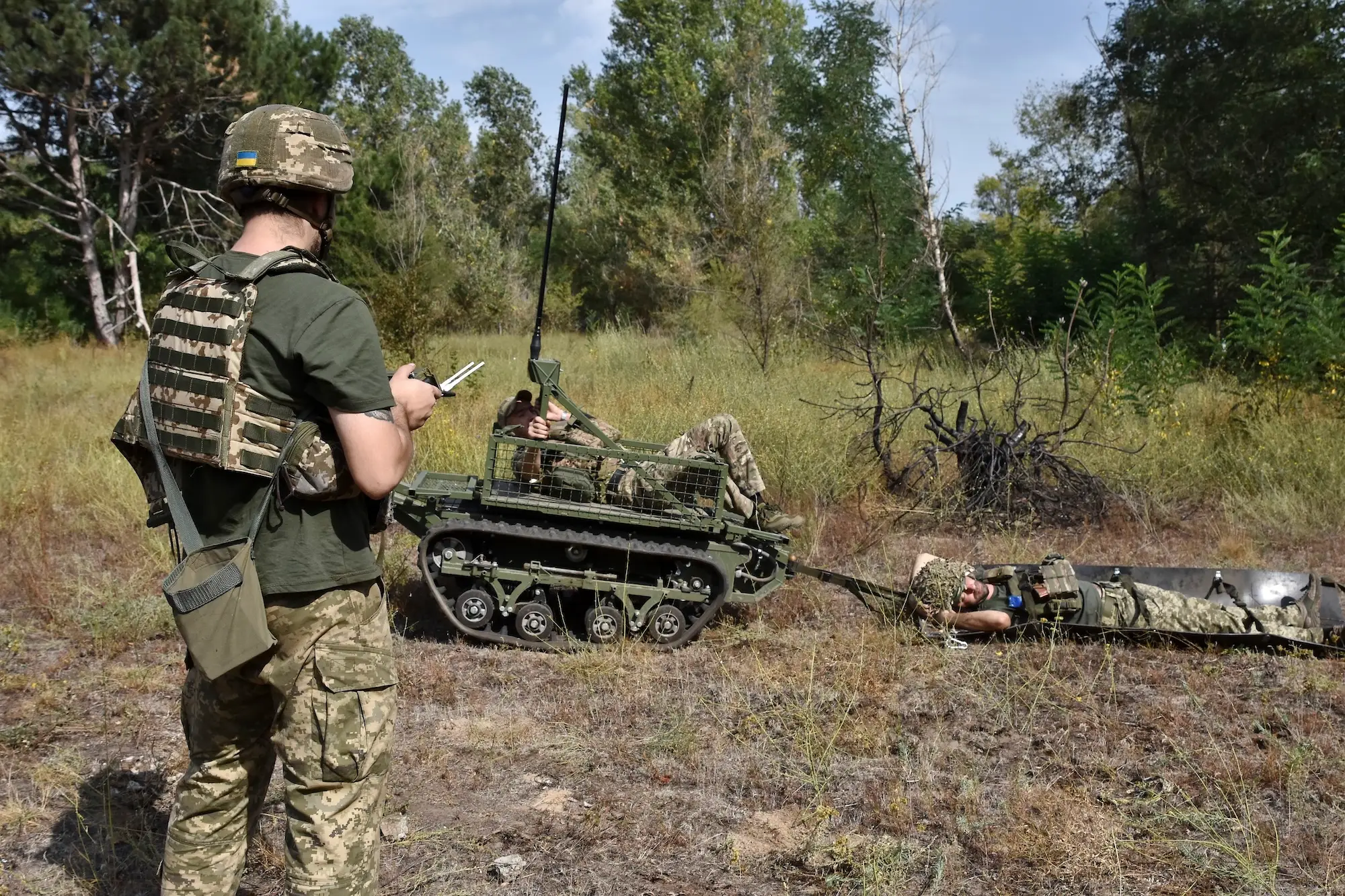 A man in camouflage gear stands watching a robot with a man lying on top pulling another man along the ground in a field