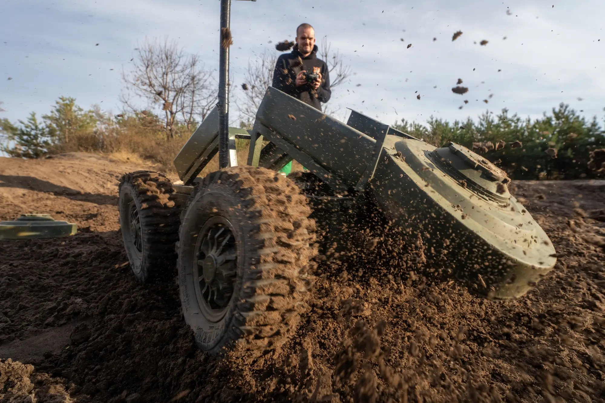 A man stands behind a robot with large wheels that is pushing a flat green mine into the earth