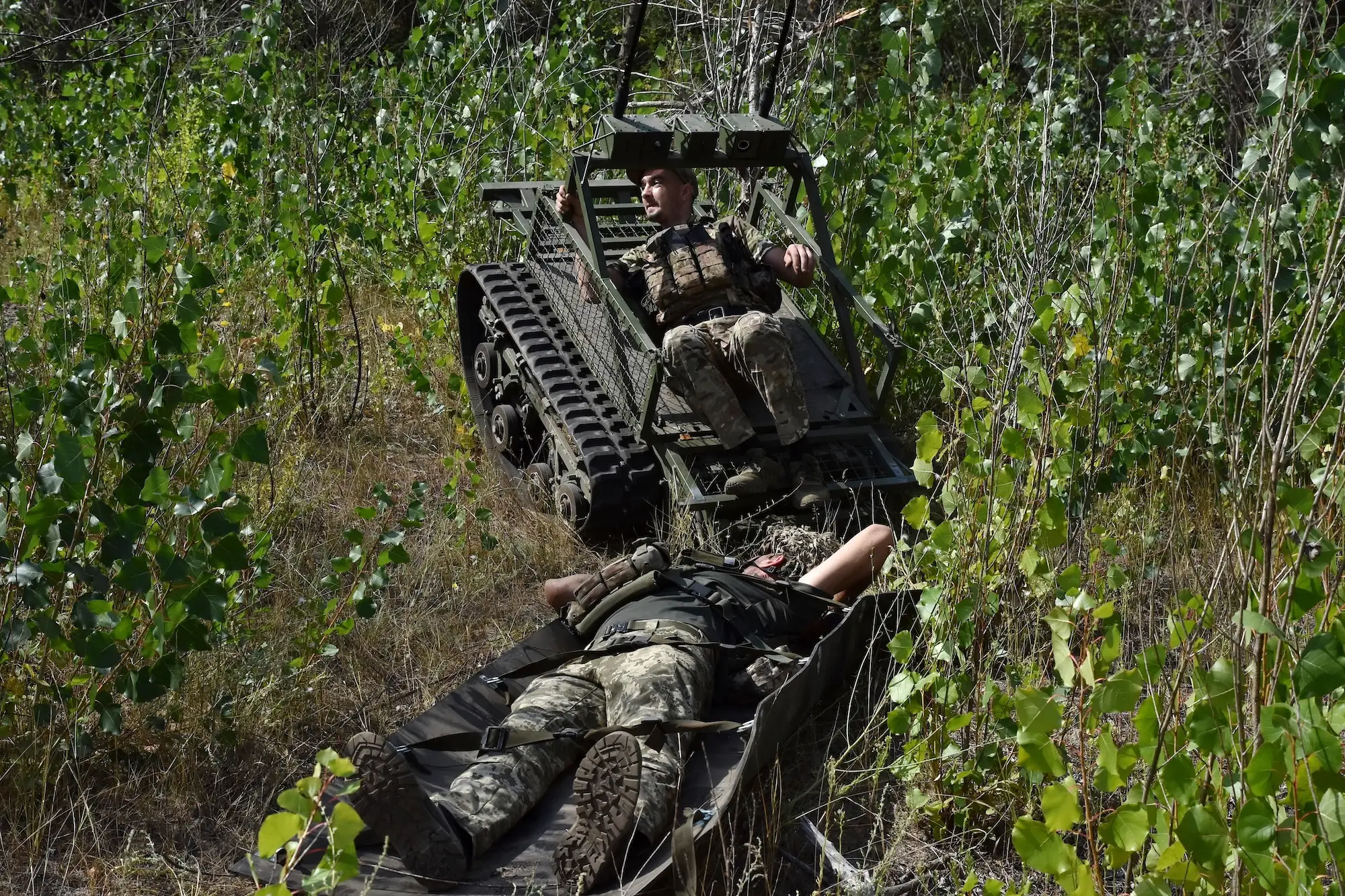 A dark green colored robot drives among foliage with a person in camouflage gear lying on top and pulling another person behind on a platform