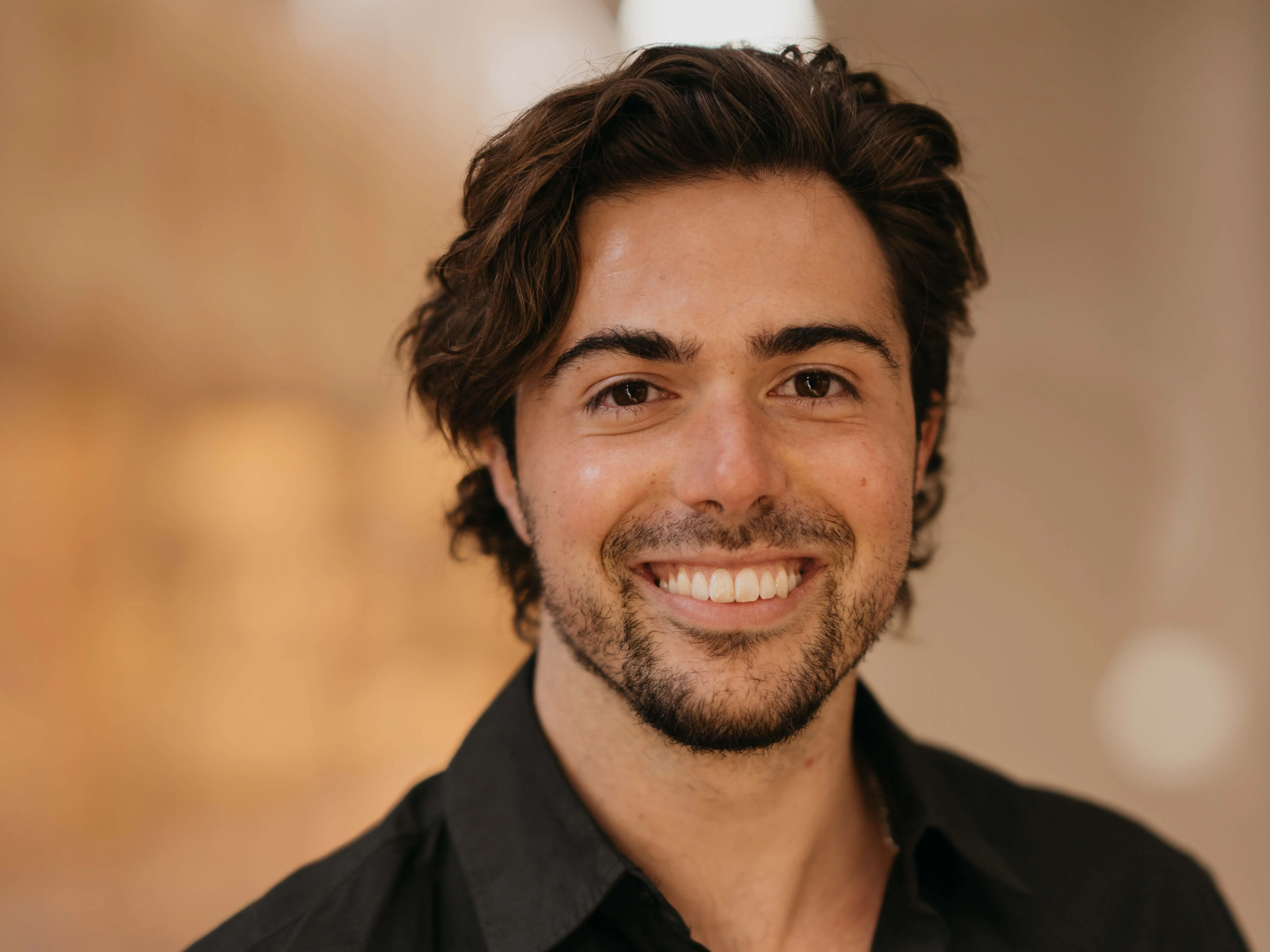 A headshot of startup founder Max Marchione smiling at the camera. He is wearing a black button-up shirt with an open collar against a background of soft-focus warm light.