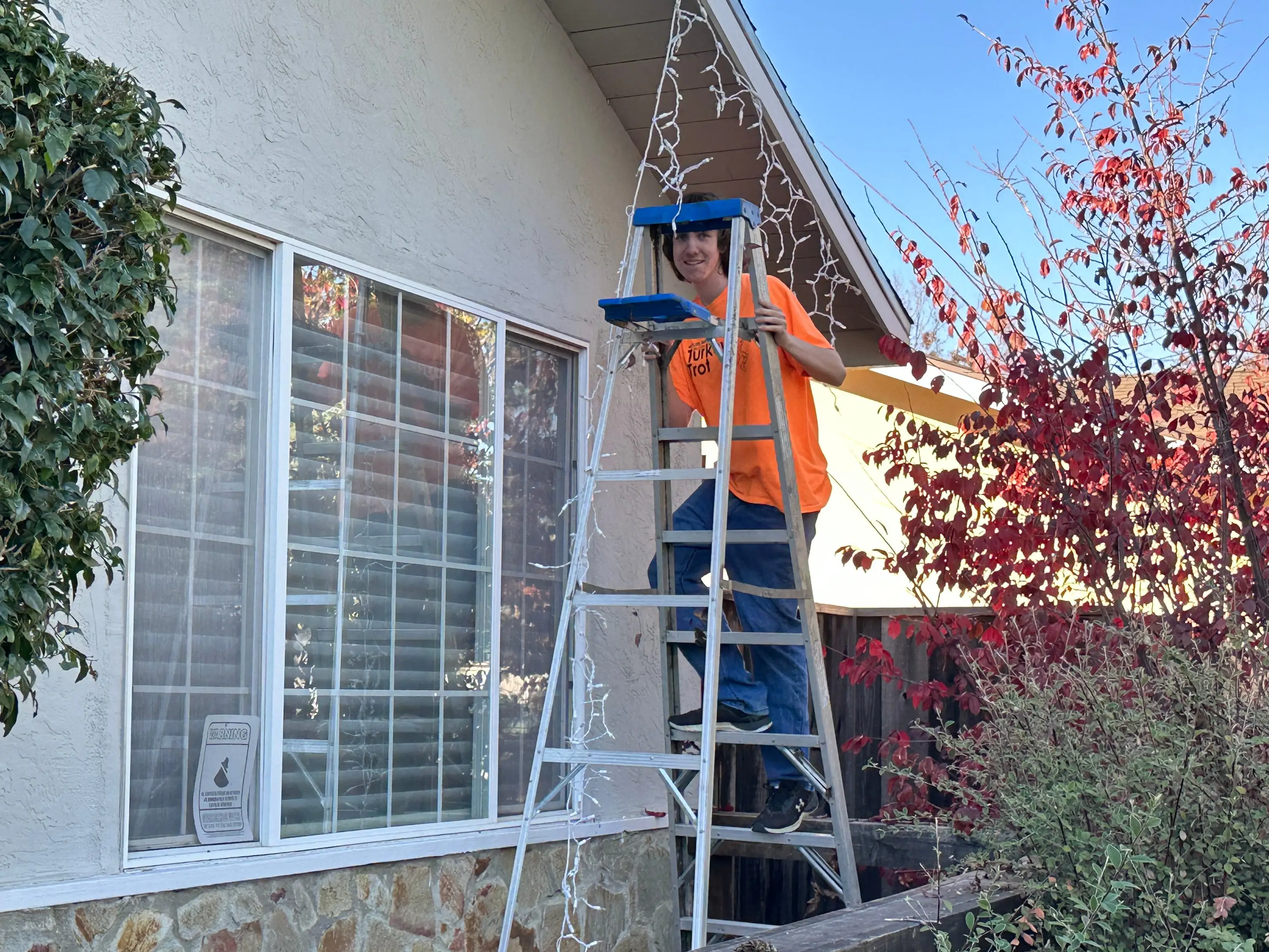 The author's son stands on a ladder while hanging holiday lights on the family home.