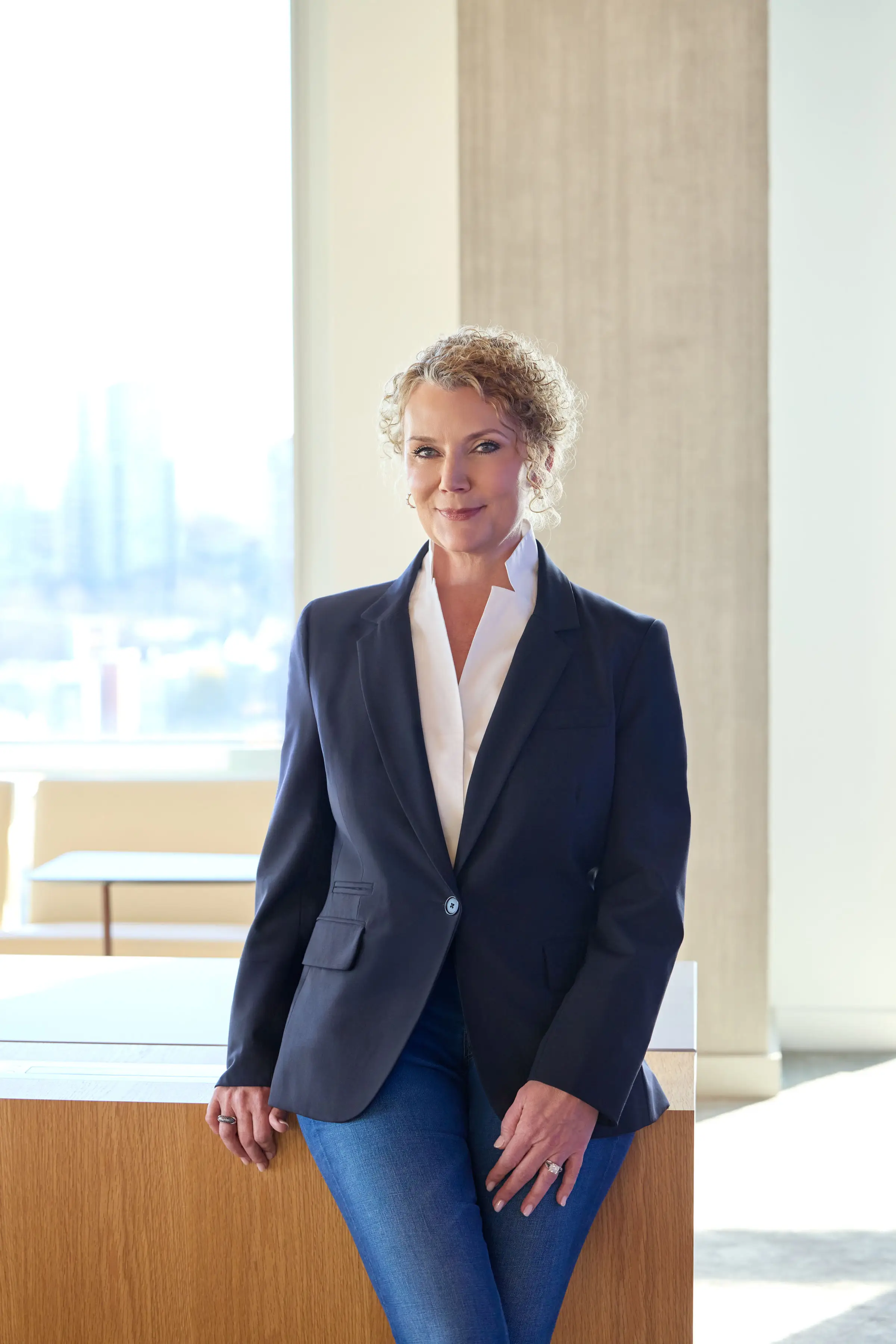 a woman in a navy blazer and jeans poses against a wooden table