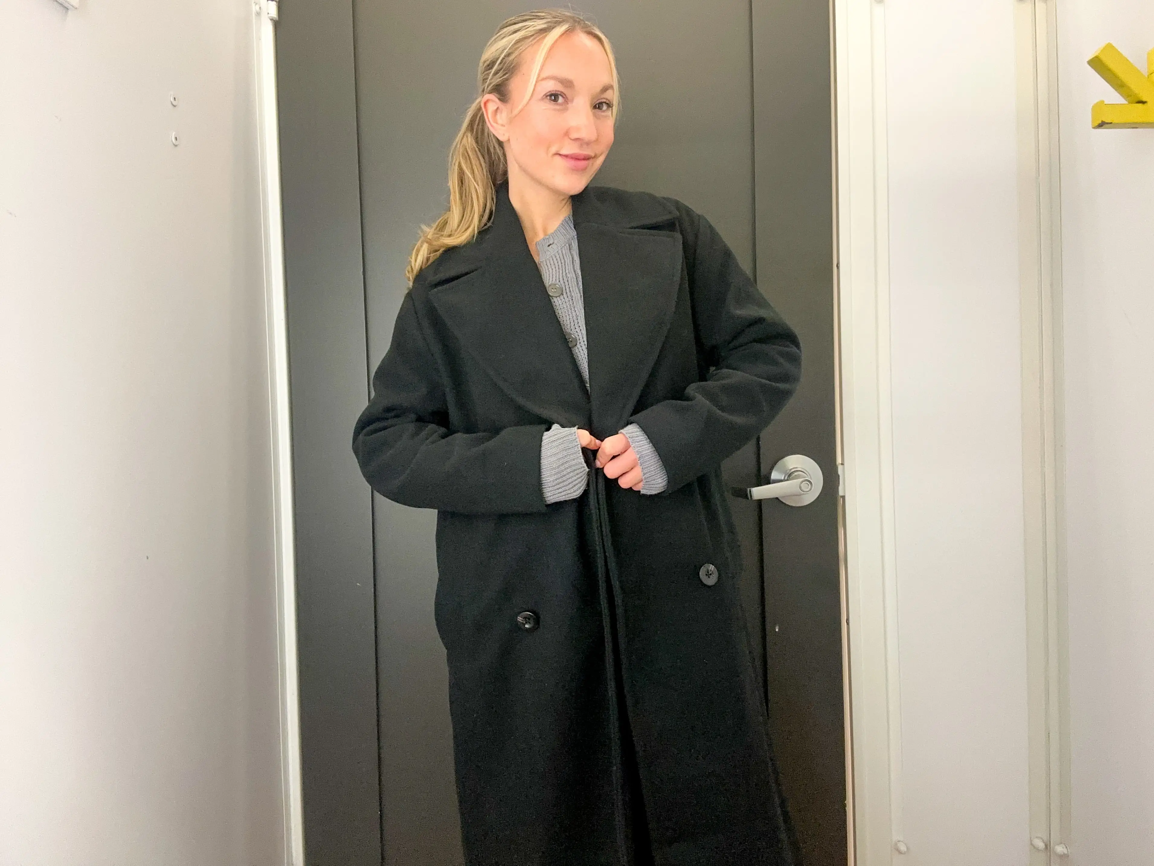 Chloe wears a long black peacoat in a fitting room.