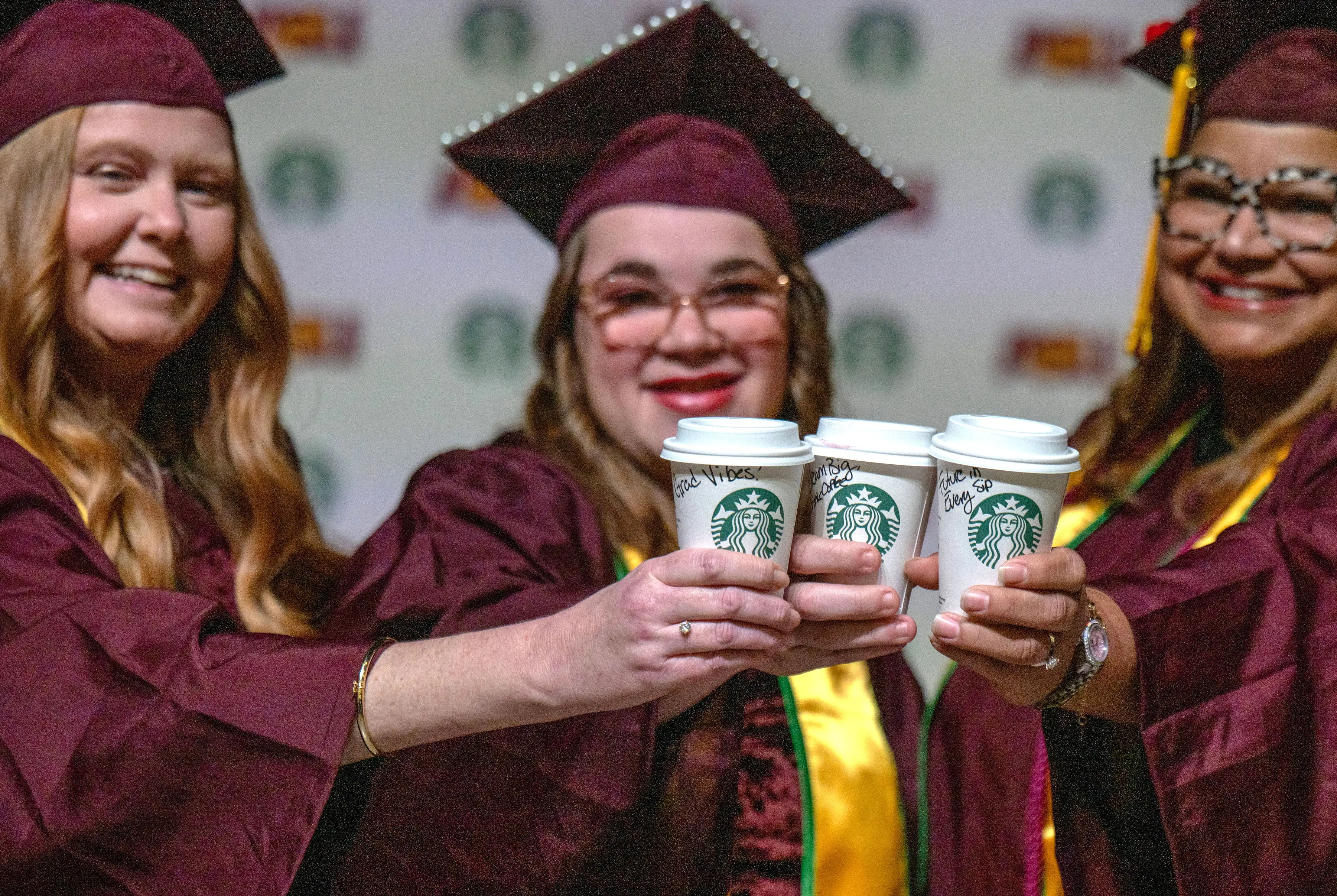 Three graduates of Starbucks' College Achievement Plan (SCAP) program cheer while holding Starbucks cups.