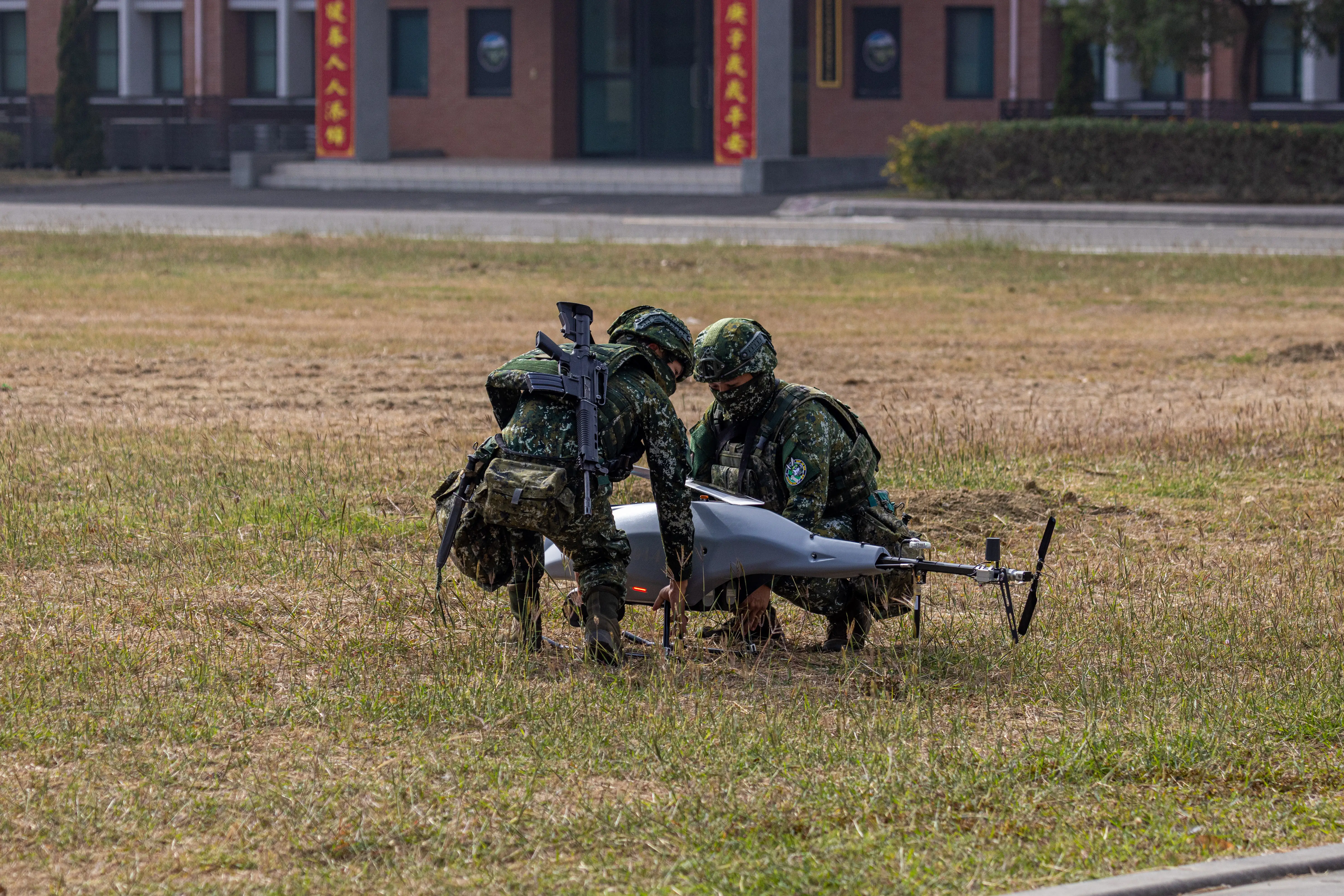 Two people wearing camouflage crouch in front of a rotary-wing drone that's sitting on a patch of greenish brown grass.