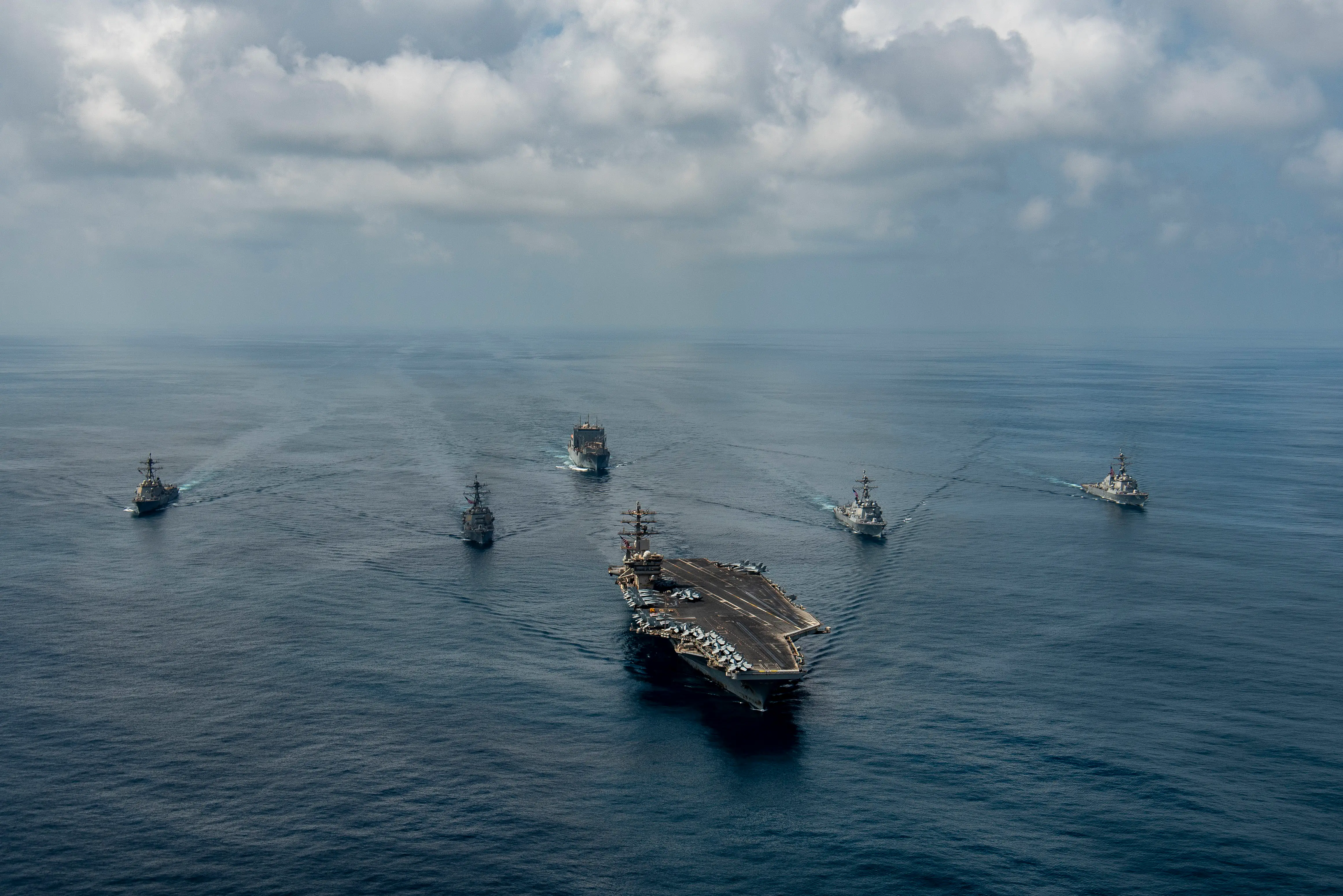 An aircraft carrier and five vessels sail in calm blue water with a cloudy sky in the background.