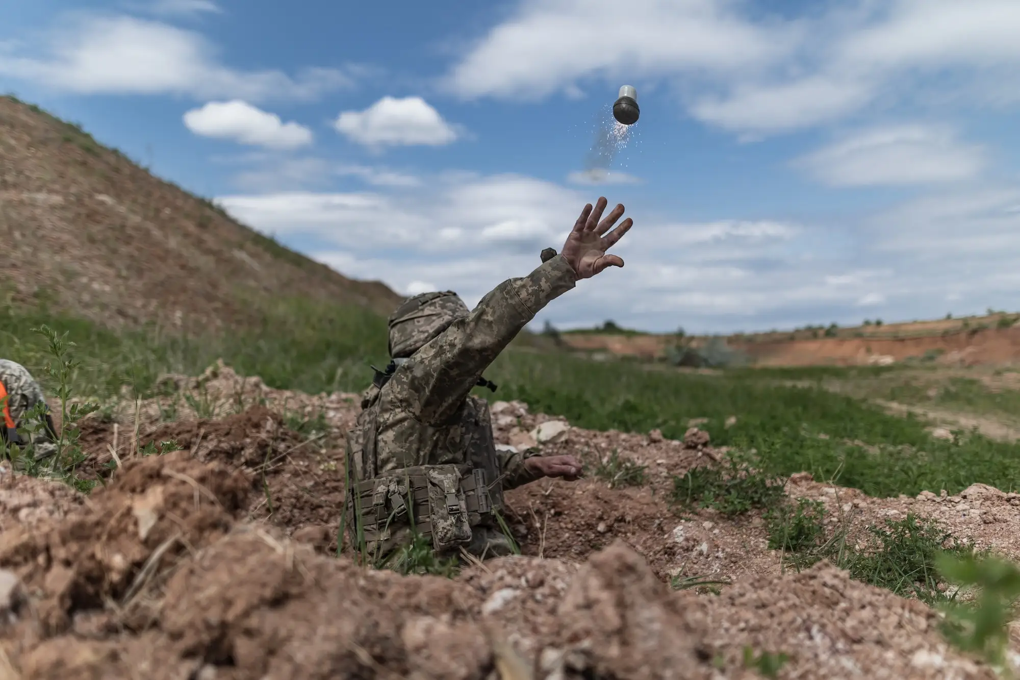 A figure wearing camouflage throws a sparking grenade into the air as they stick out from the ground under a blue sky