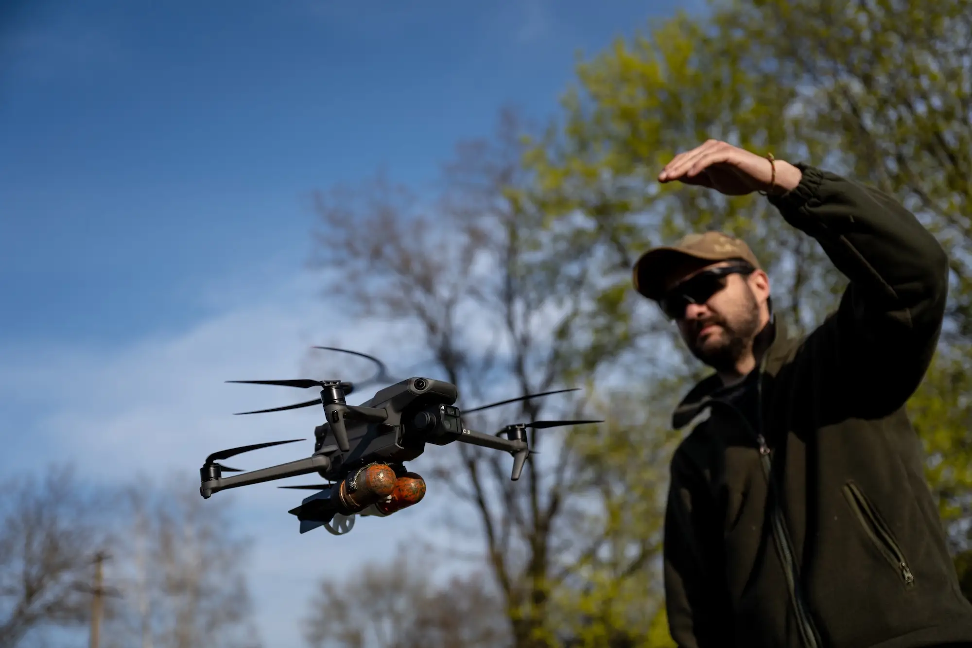 A man in a dark fleece and cap stands with his arm up beside a small black drone that's hovering in the air with two red grenades attatched to it.