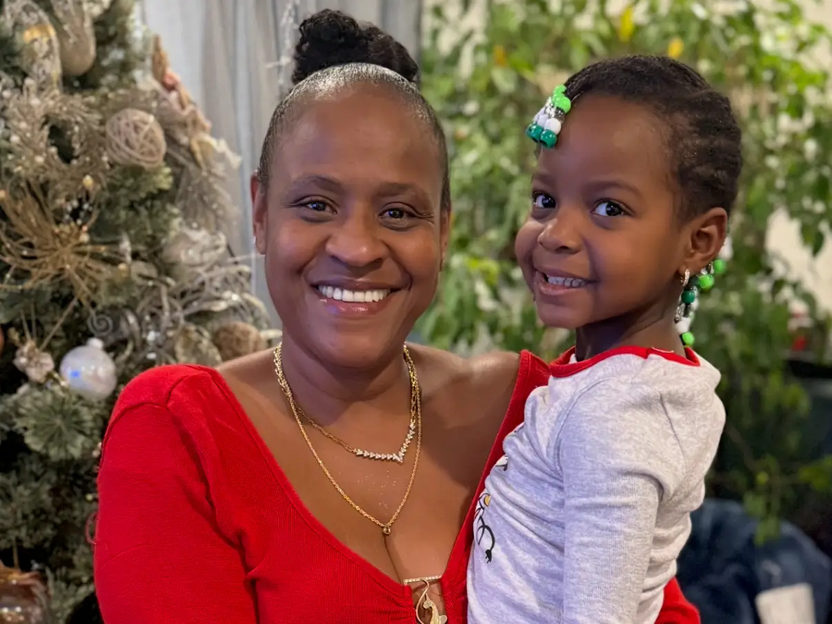 A mom and her little daughter are standing next to a Christmas tree.