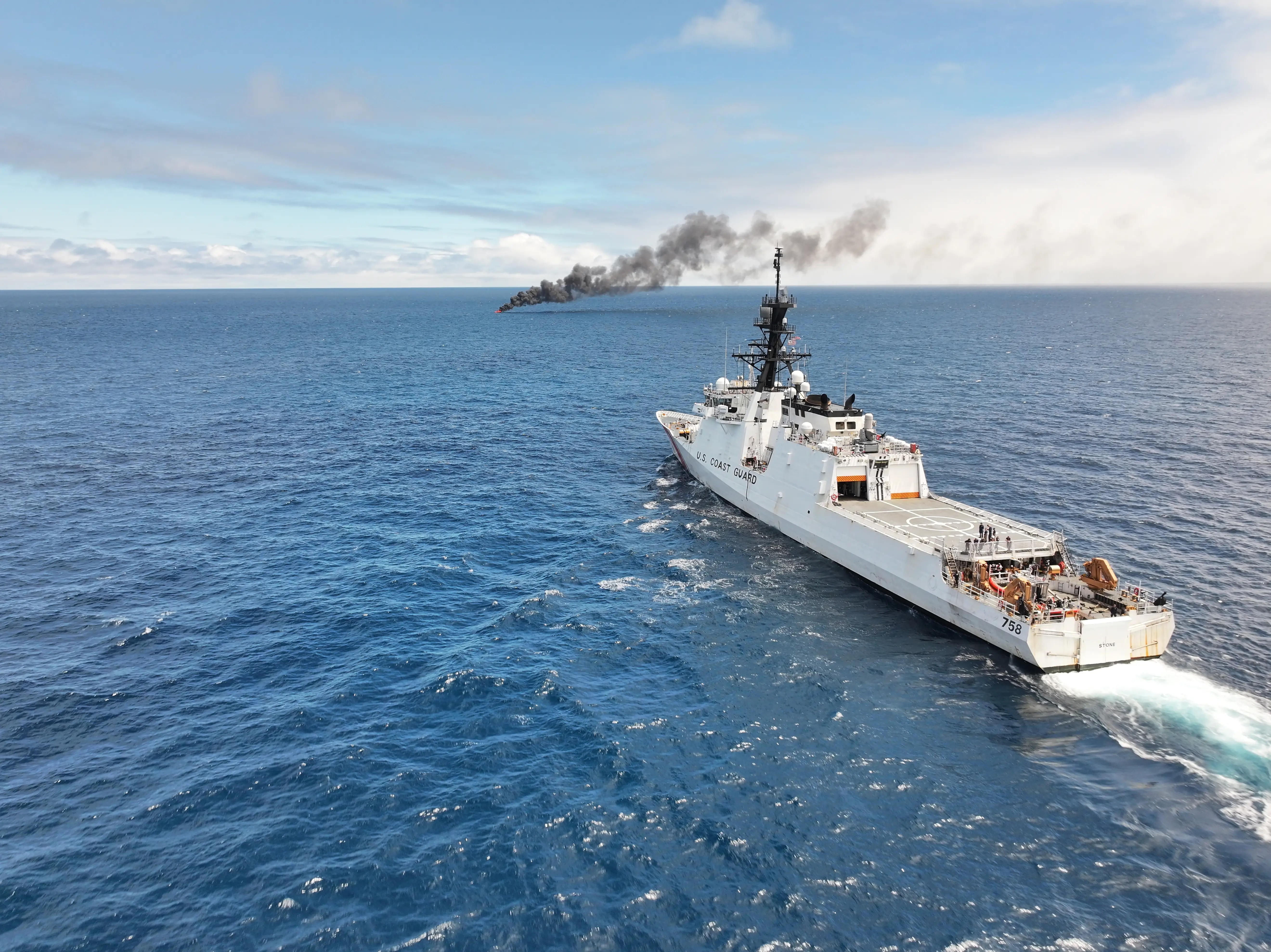A large white vessel waits in blue water with a plume of smoke rising from the water in the distance. The sky is cloudy.