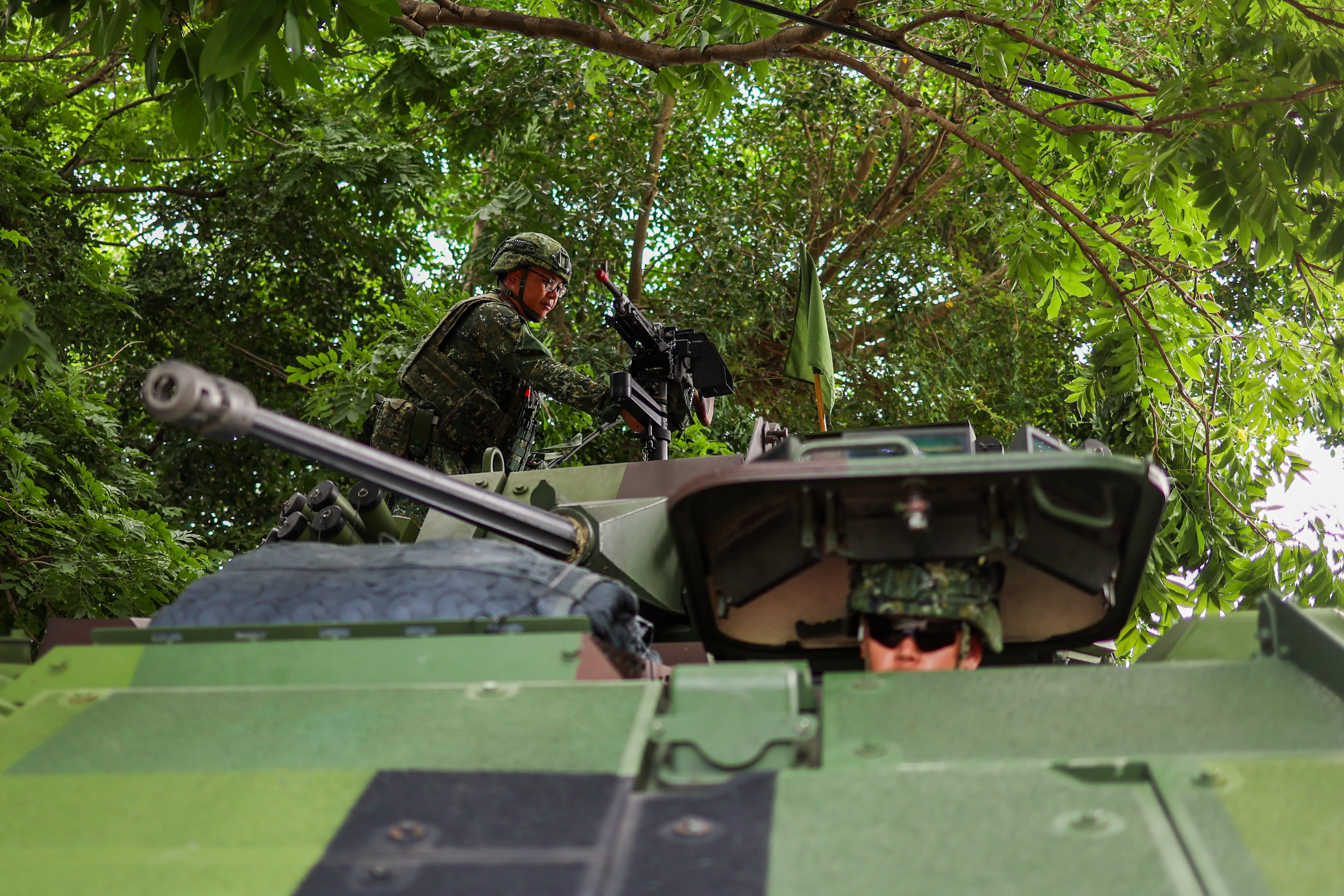 Two Taiwanese soldiers operate a tank, which is seen close-up, with a line of green trees and leaves in the background.