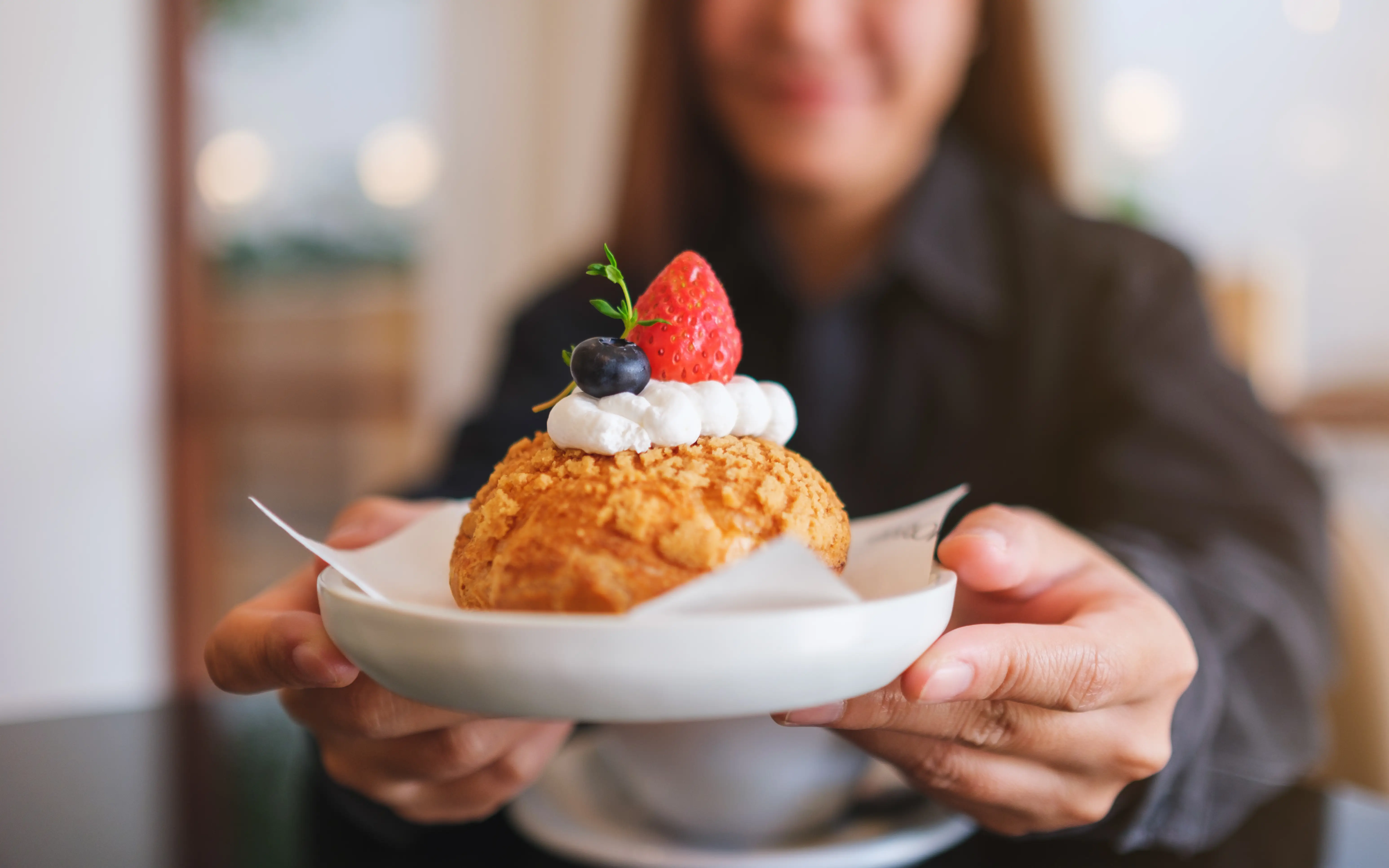 A woman in soft focus holding a fruit and cream pastry treat toward the camera.