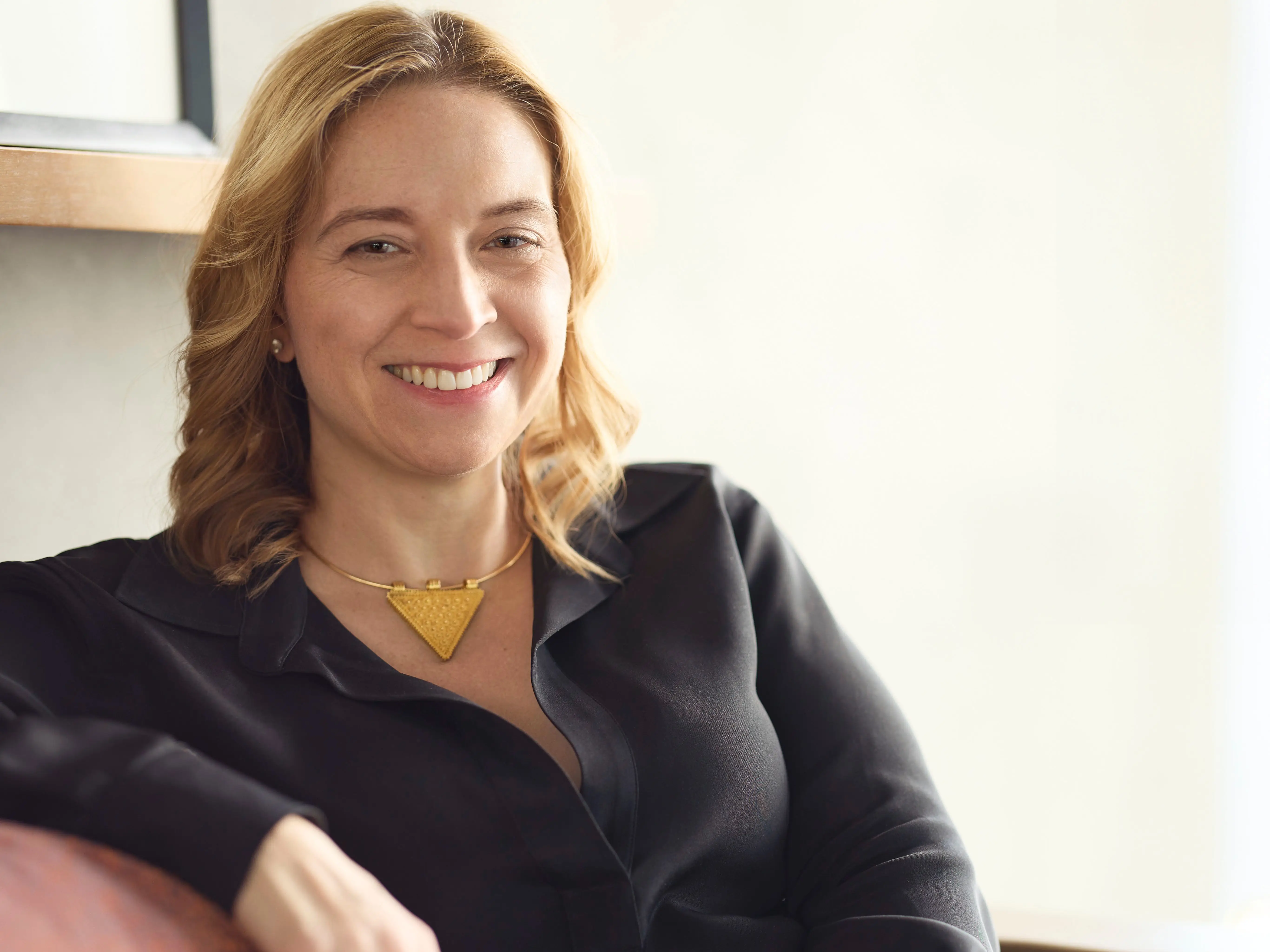 Alicia Pittman sitting on a couch in black outfit and gold-colored necklace.
