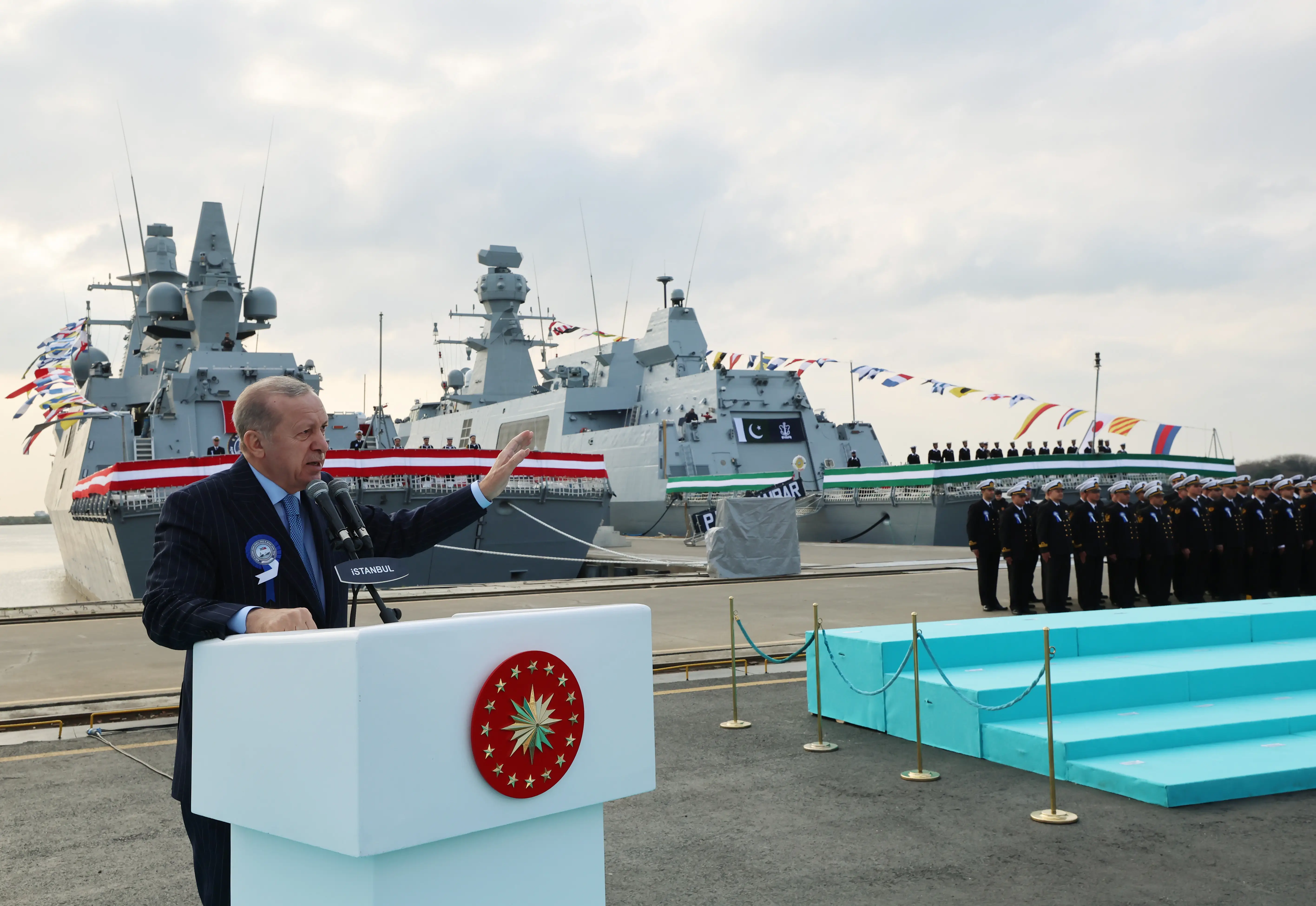 Turkish President Recep Tayyip Erdogan speaks in front of two warships at a shipyard.