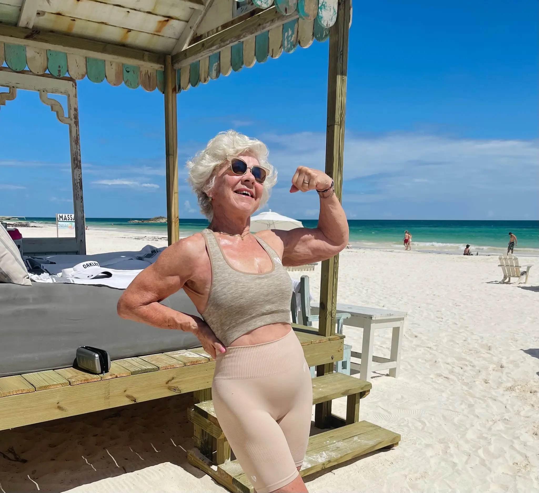 An older woman flexes her muscles on a beach.