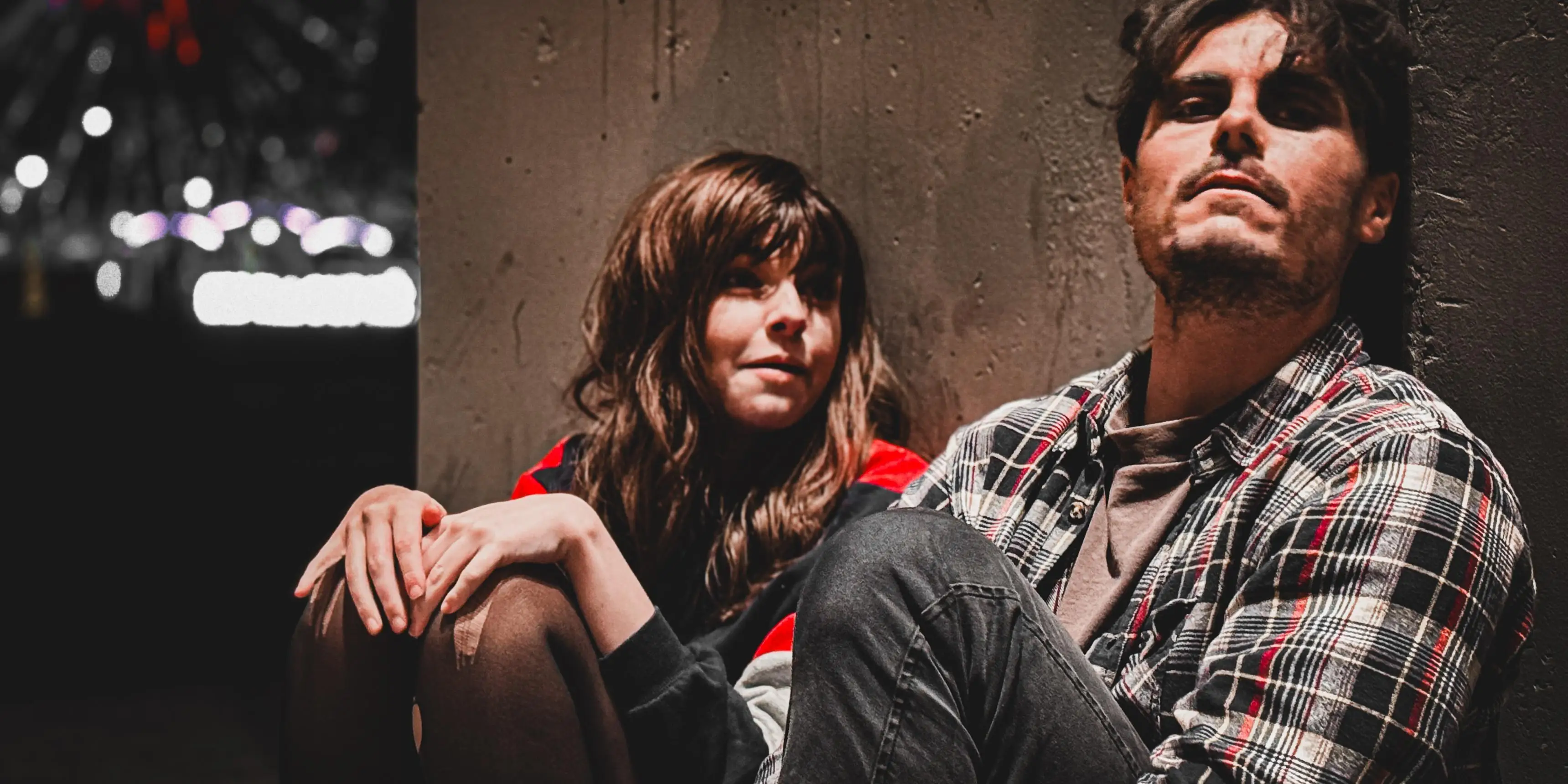 Two people sitting down, one woman on left with ripped tights and greasy hair. On right, a man with similarly dirty look.