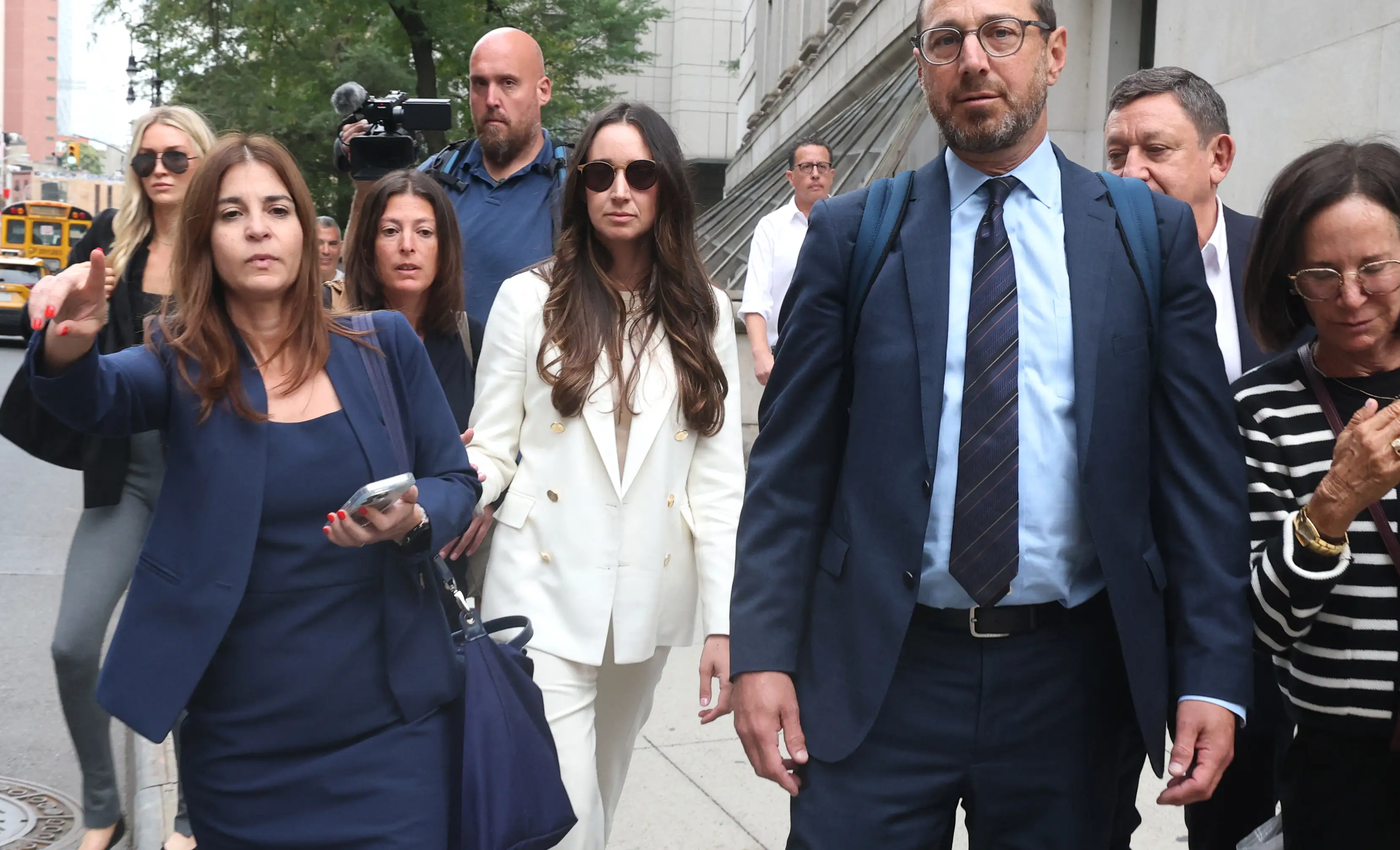 Charlie Javice, with brown hair, sunglasses and a white pantsuit, stands in the middle of a group of men and women in suits as she walks down a sunlit Manhattan sidewalk outside of a courthouse. A TV camera is visible behind her.