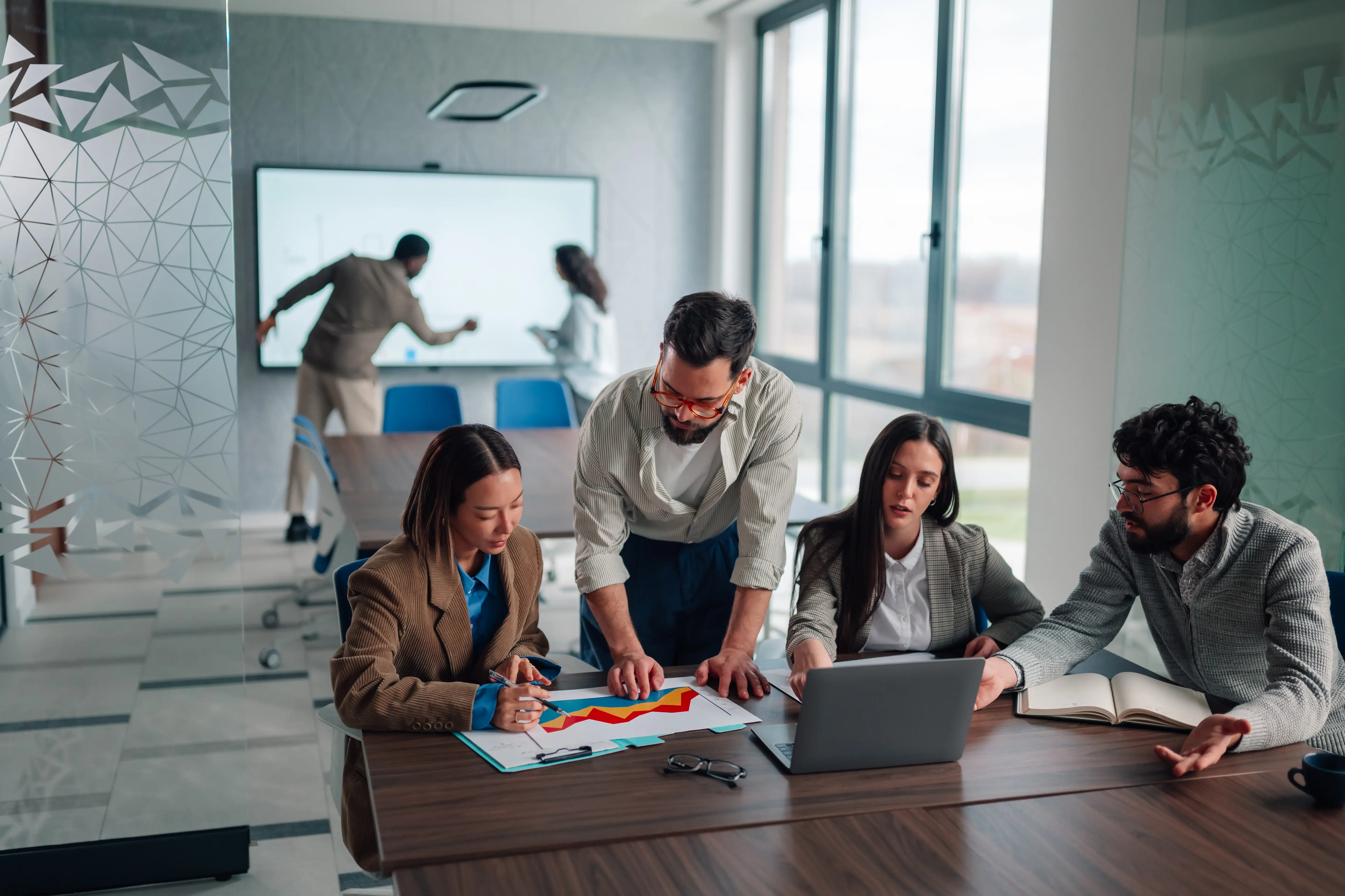 Coworkers sort through a chart and a computer while colleagues work on a smart board in the background.