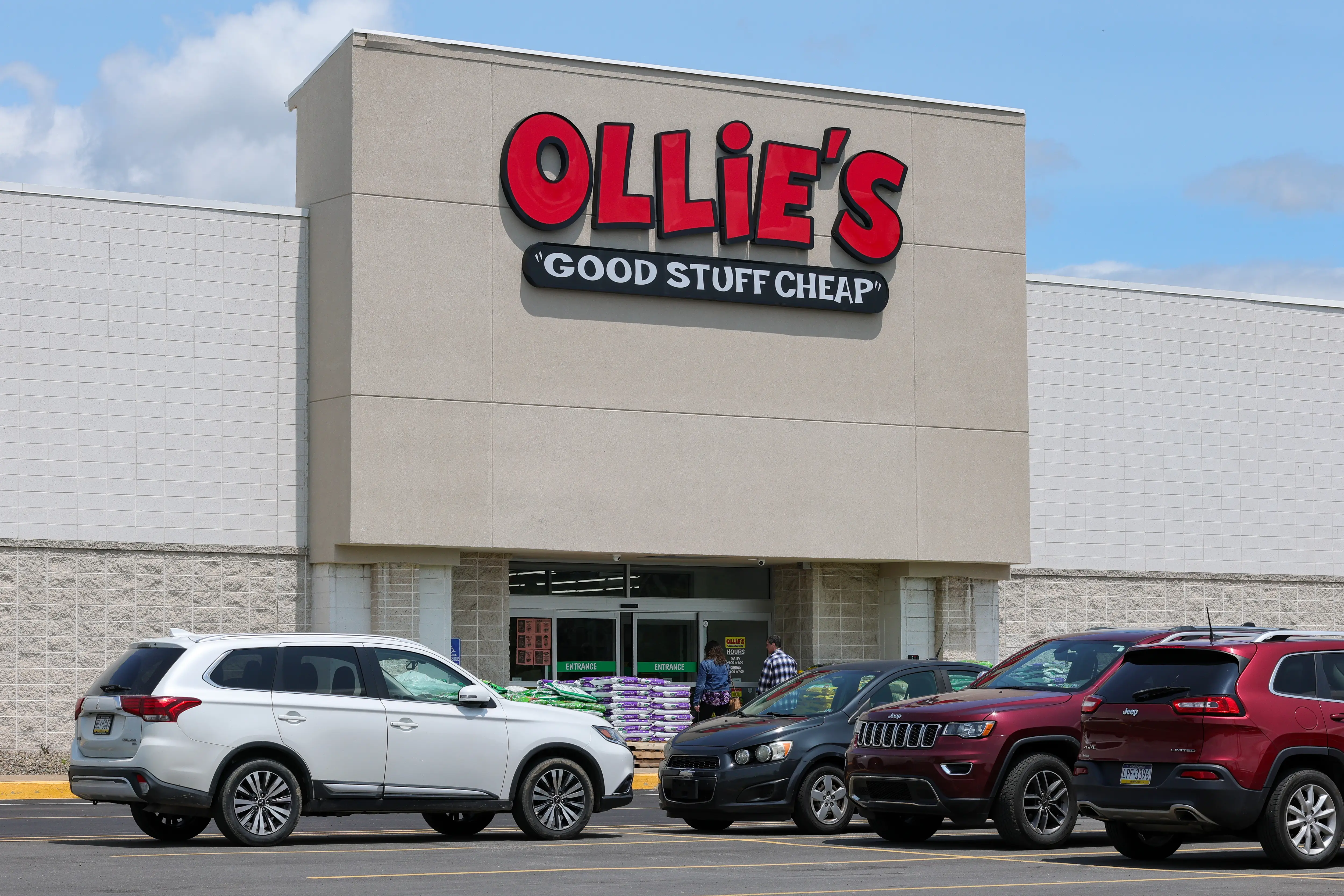 Cars sit parked outside of an Ollie's Bargain Outlet store as customers walk in and potting soil sits in bags outside of the front doors. Above the entrance, a red-and-black sign reads: 