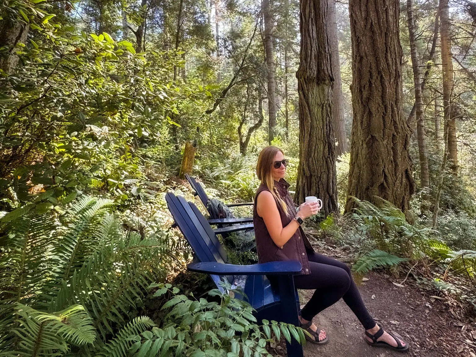Emily holds a cup of coffee while sitting in a chair under large trees.