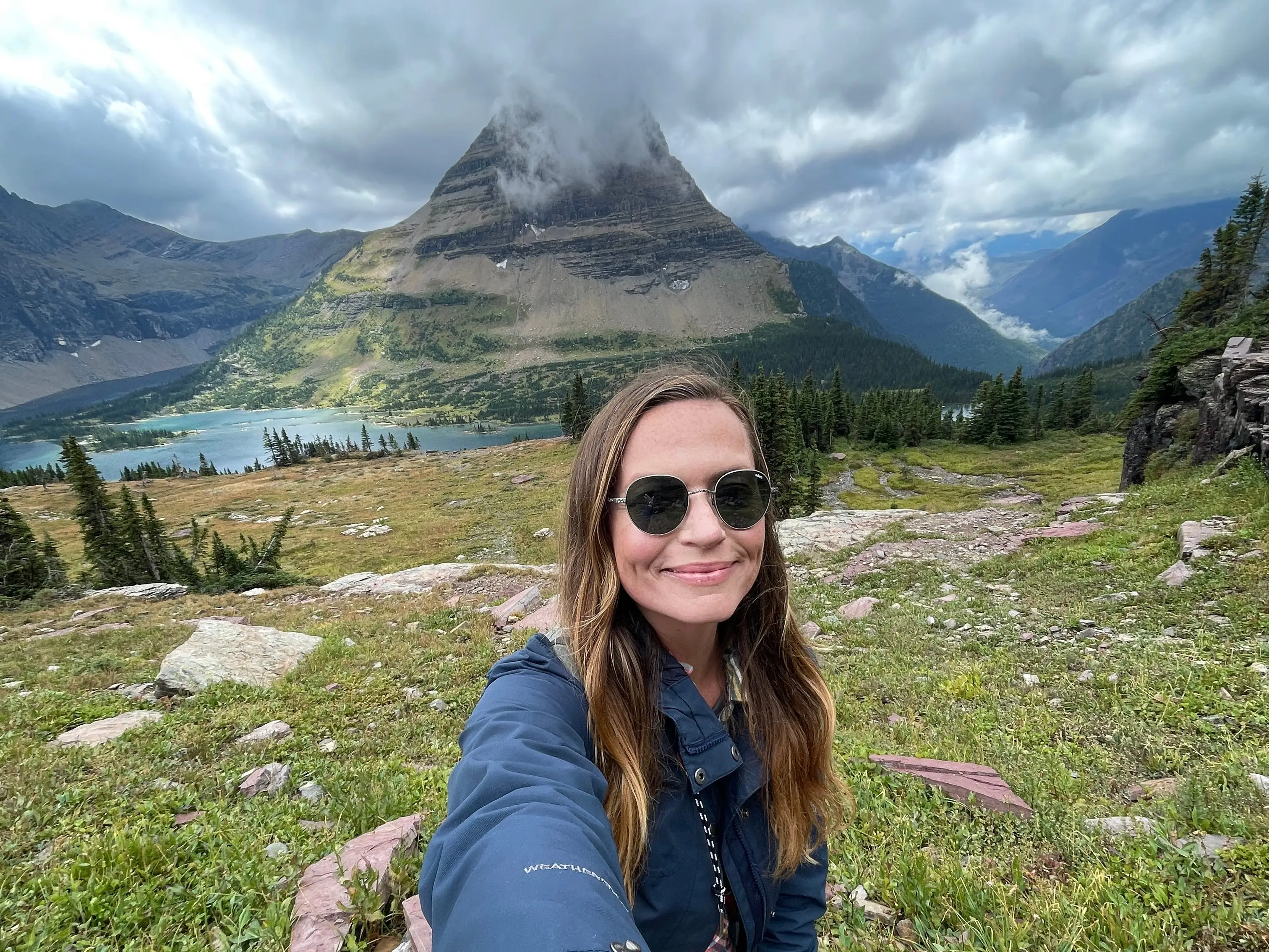 Emily takes a selfie in front of a mountain surrounded by a lake on a cloudy day.