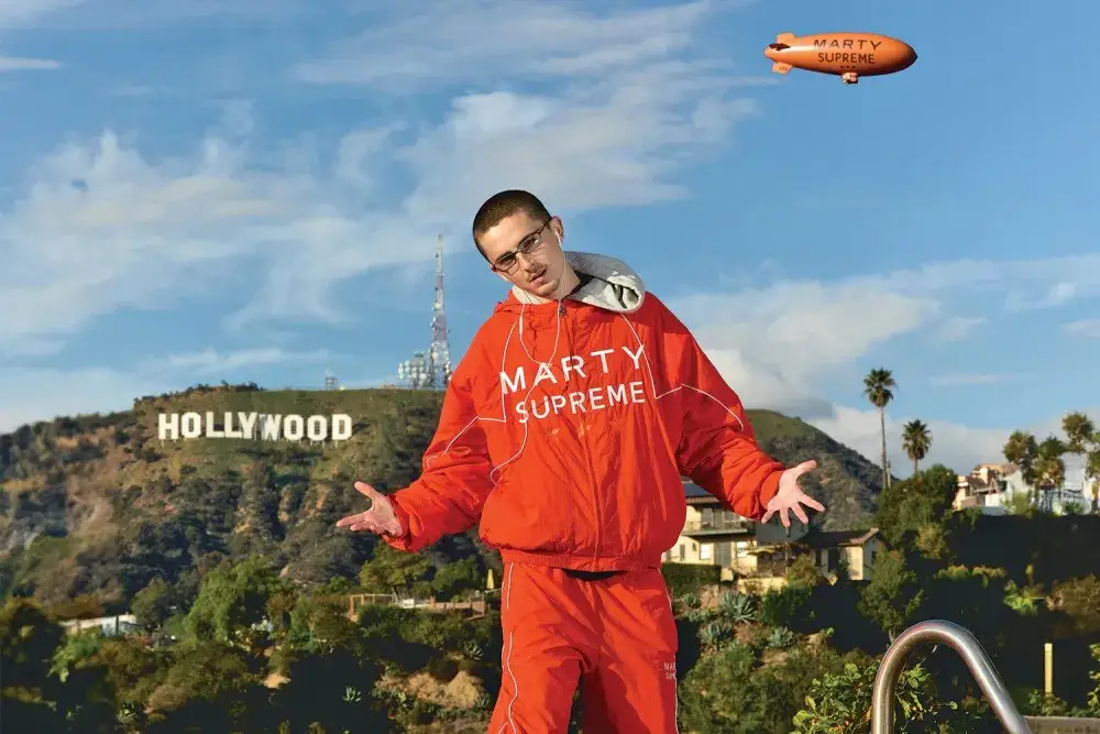 Timothee Chalamet in a Marty Supreme jacket in front of the Hollywood sign with a blimp in the sky