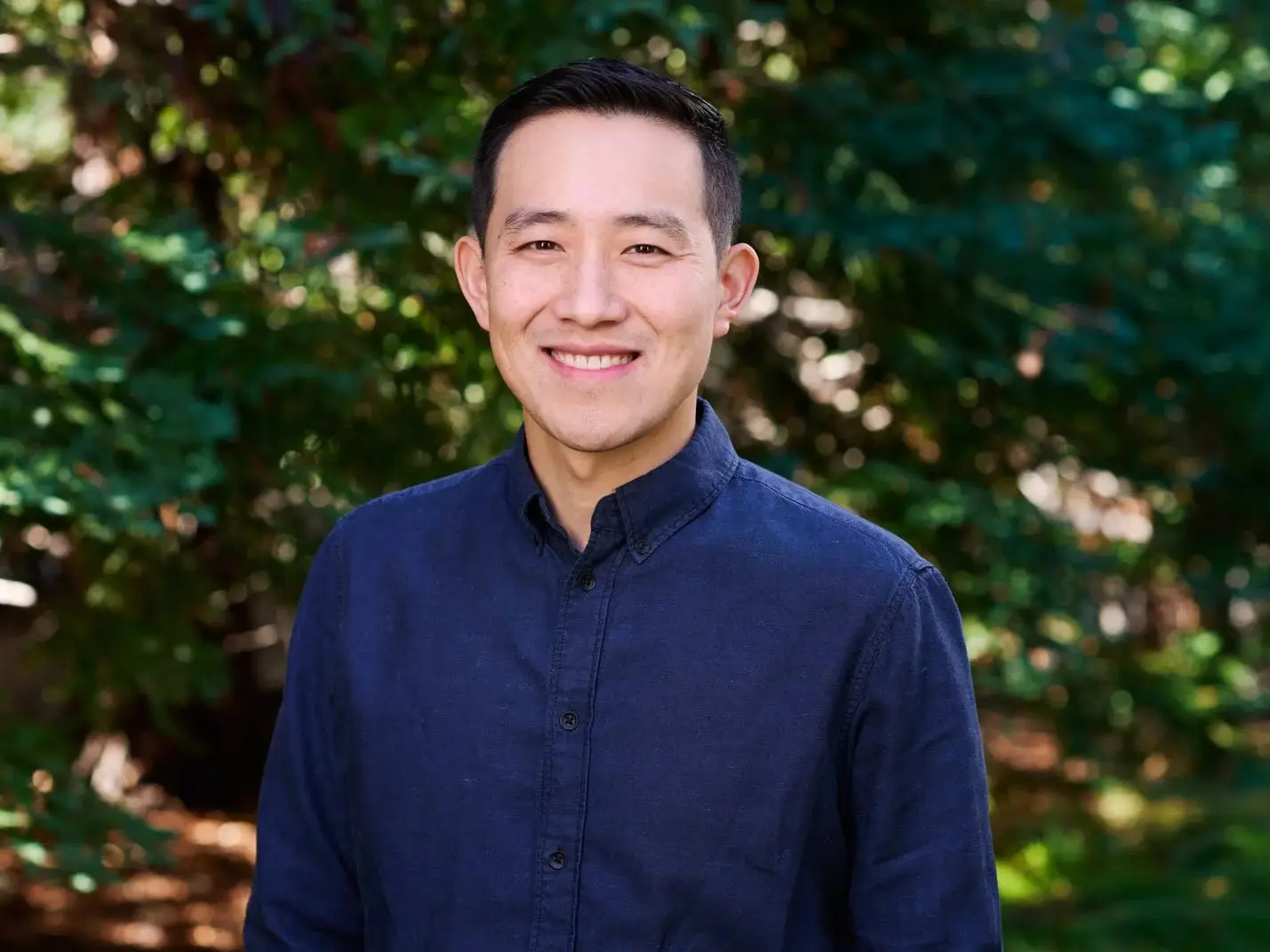 A close-up of a man smiling for a portrait, standing in front of a wooded area.