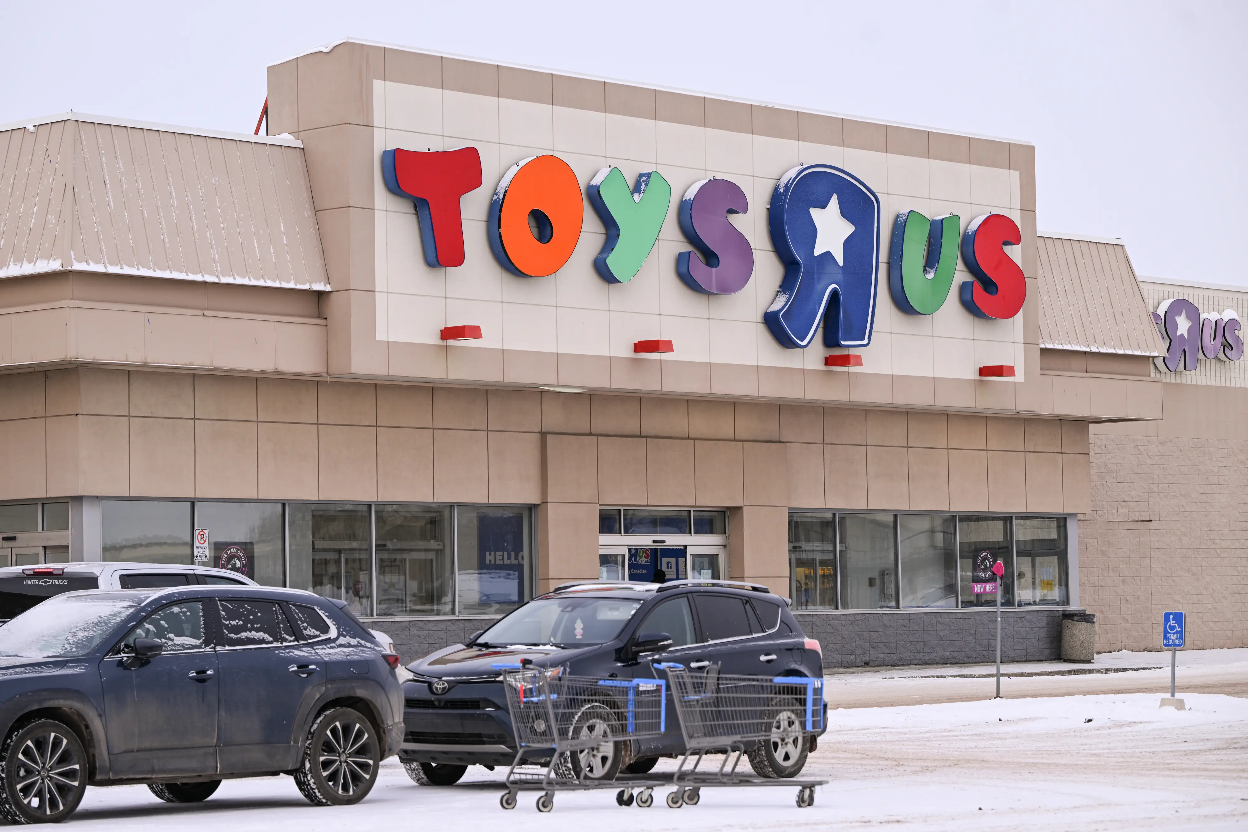 The entrance to a Toys R Us store with a lighted sign above and dark-colored SUVs and some shopping carts in the parking lot.