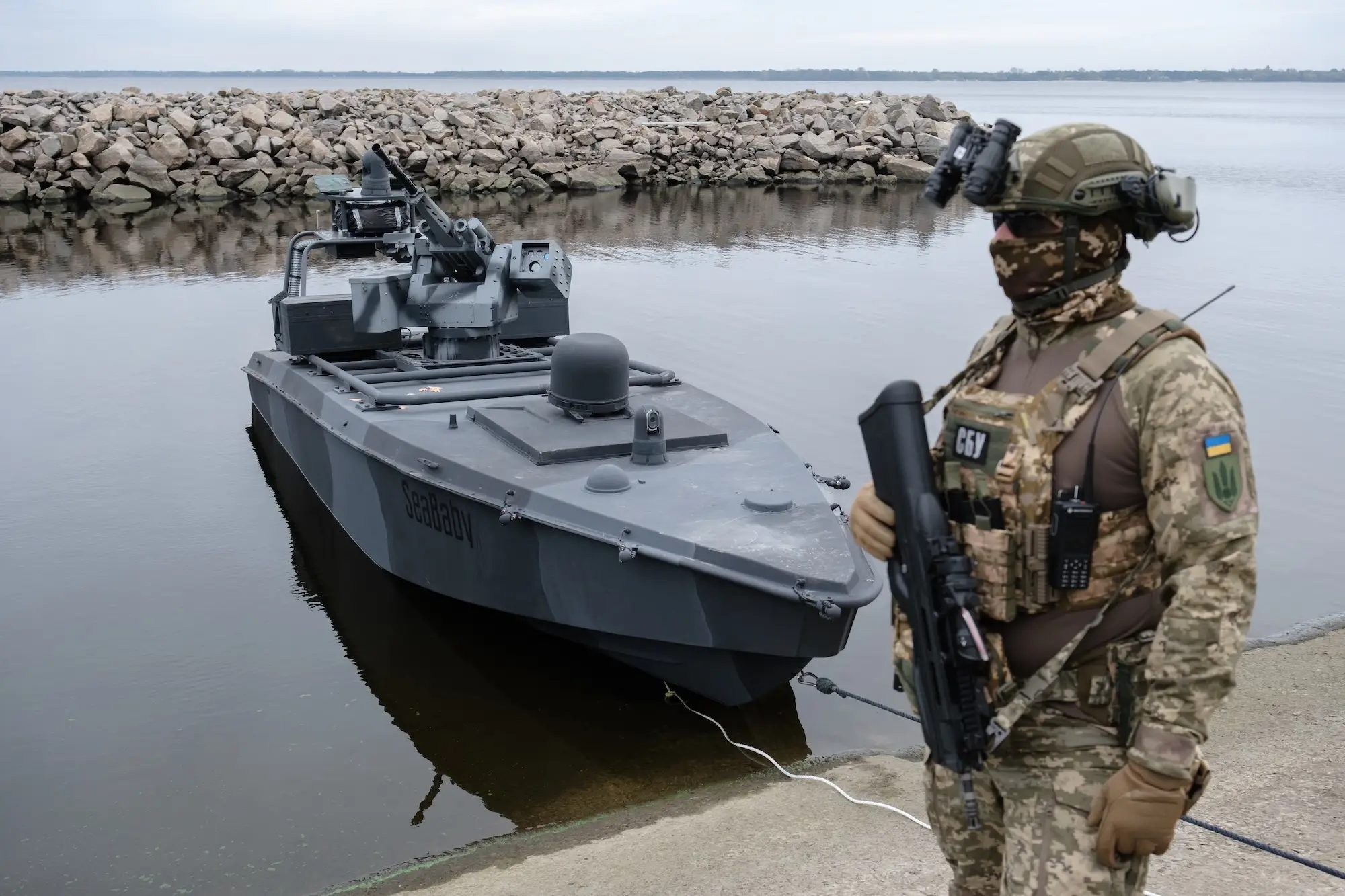 A large grey unmanned naval drone in still water with a person in camouflage gear standing on concrete beside it