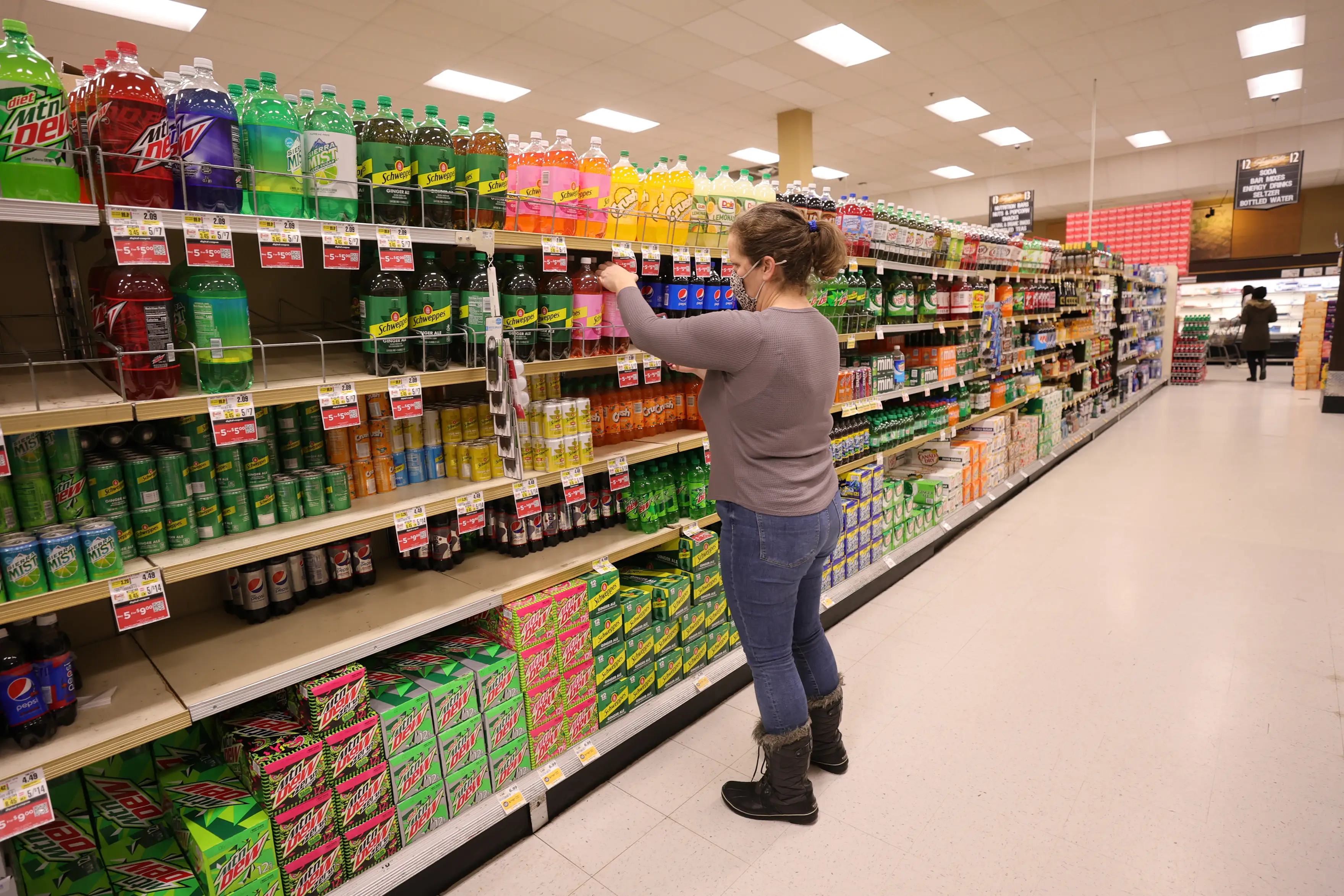 A woman wearing a face mask reaches for a two-liter bottle of soda on a grocery store shelf.