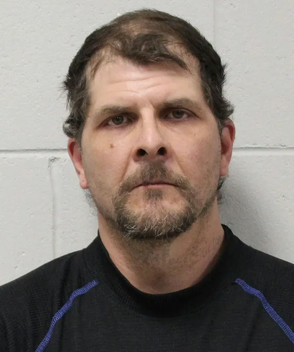 Mugshot of a man with dark hair, a beard, and a mustache, wearing a black shirt with blue stitching.