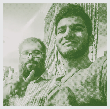 candid photo of bearded man in glasses in background holding up two fingers and younger man smiling in foreground